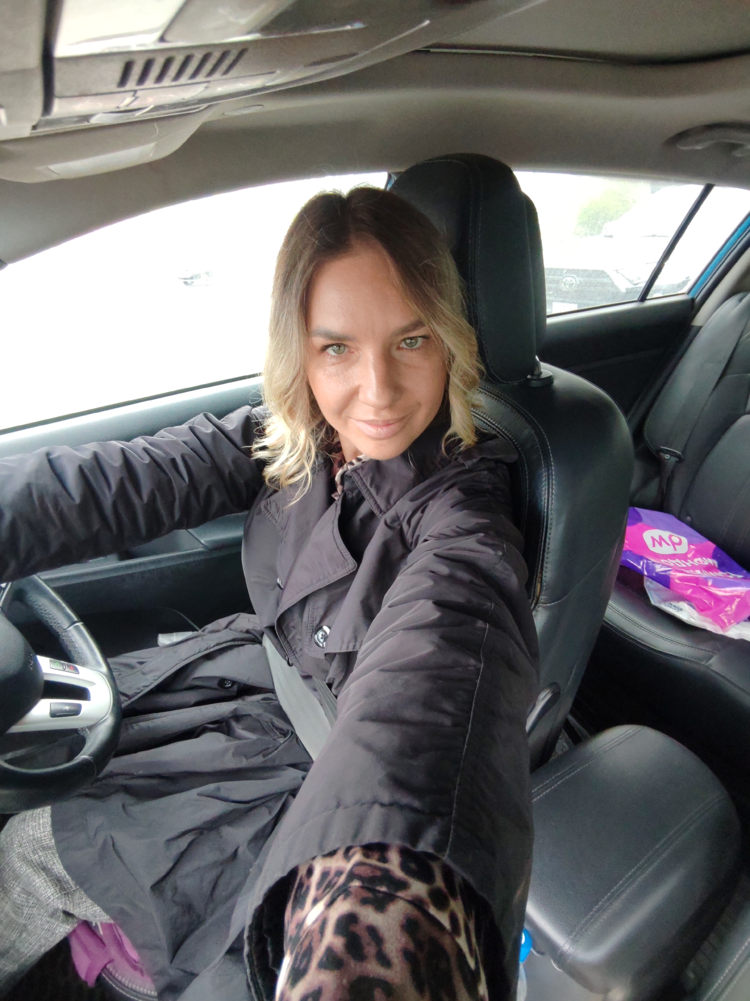 A woman is captured in a selfie taken from the driver's seat of a car. She has blonde-brown hair, green eyes, and a slight smile, looking directly at the camera. She is wearing a dark, possibly black or charcoal gray, trench coat or jacket. A leopard print pattern is visible on the sleeve of a garment worn underneath her coat on her right arm. A seatbelt is fastened across her chest.
The car's interior is visible, featuring black leather seats, a multi-function steering wheel with silver accents, and the car's roof liner. Through the car windows, an overcast sky and the outlines of other vehicles suggest it is daytime with possibly cloudy or rainy weather.
On the rear passenger seat, a purple plastic bag is present. White text is printed on the bag, with the letters "mp" being clearly discernible. The overall scene suggests the woman is taking a casual selfie while parked or waiting in her car.