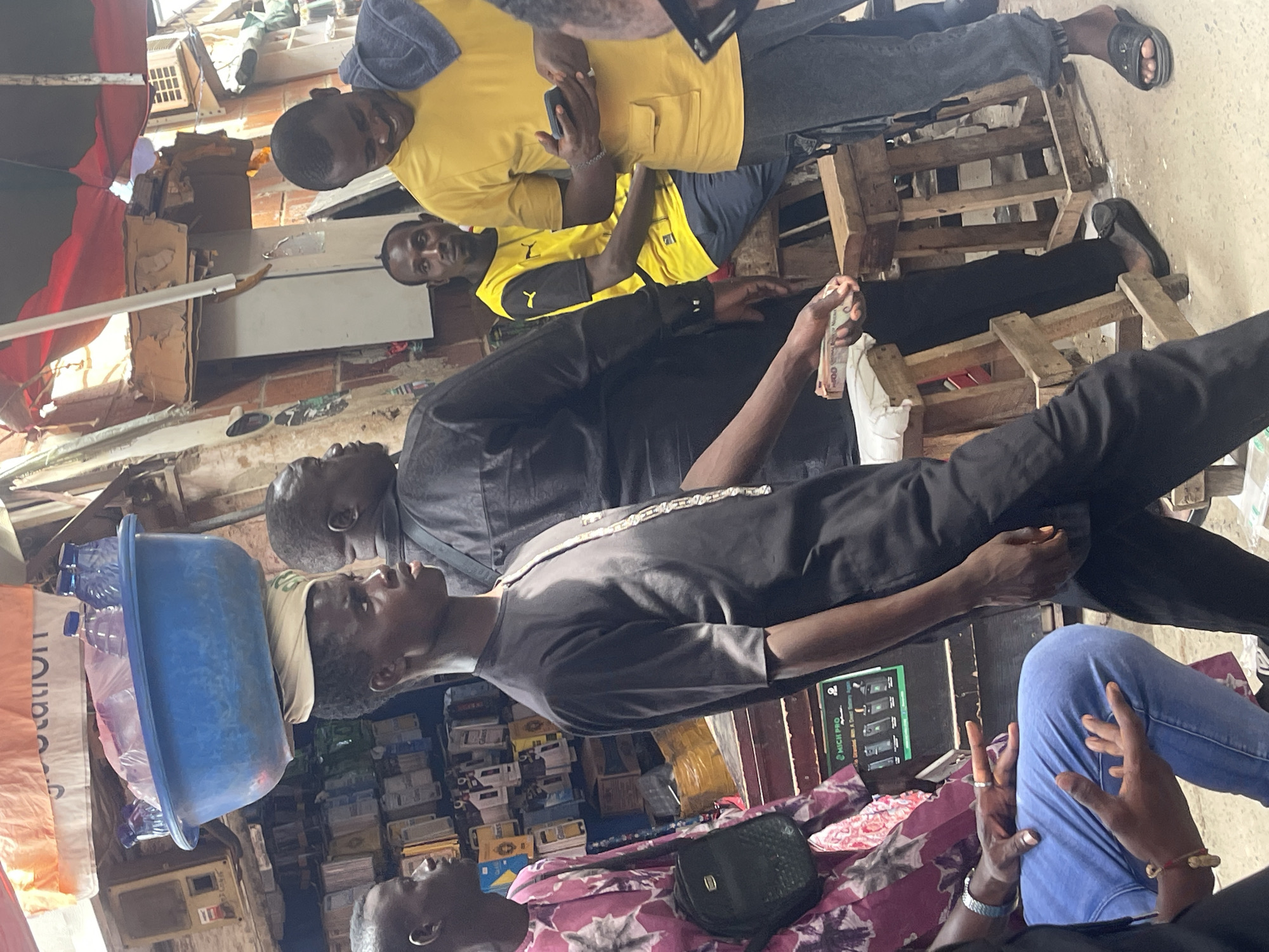 The image captures a group of men gathered in what appears to be an outdoor or semi-outdoor market stall, likely in Abuja, Nigeria. The setting features makeshift wooden structures, possibly pallets or crates, used for seating and display.
Several men are present. In the upper part of the image, two men are visible: one wearing a bright yellow t-shirt with a dark blue item on his head, holding a mobile phone; next to him, another man wears a yellow and black Puma branded t-shirt. Another man in a black long-sleeved shirt is seen holding a stack of what appears to be Nigerian Naira notes (500 or 1000 denominations are visible). Further down, a man in a dark grey t-shirt and a light-colored cap stands. In the bottom right, parts of other individuals are visible, including hands (one with rings and a bracelet) belonging to someone wearing blue jeans, and a man in a patterned, possibly pink or red, shirt with a black handbag on his lap.
The background shows various packaged goods, boxes, and other items stacked, suggesting a retail environment. A blue plastic basin containing plastic bottles is visible on the left. Brick walls and some overhead structures are also part of the scene.
The activity suggests a casual gathering or business interaction. The man holding money might be involved in a transaction or showing it to others. The men generally appear relaxed. The scene is brightly lit, indicating daytime.
Visible text includes the "Puma" logo on one shirt, "MICK PRO" on a green sign, and "station" on a white and orange bag.