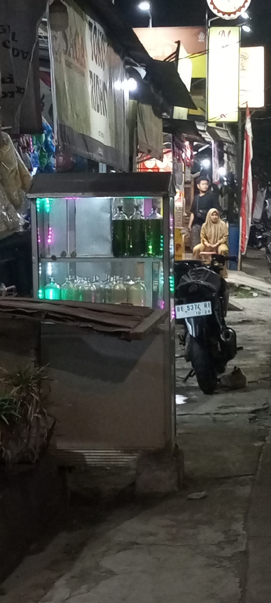In Setu, Indonesia, at night, a street vendor's stall features a glass display case filled with bottles of green liquid, illuminated by colorful LED lights. A motorcycle with license plate BE 5374 RI is parked nearby. Two people are visible behind the stall: a man in a black shirt and a woman wearing a tan hijab and clothing, seated on a stool. The background shows a dimly lit street with signs and more stalls, suggesting a bustling market or food street.