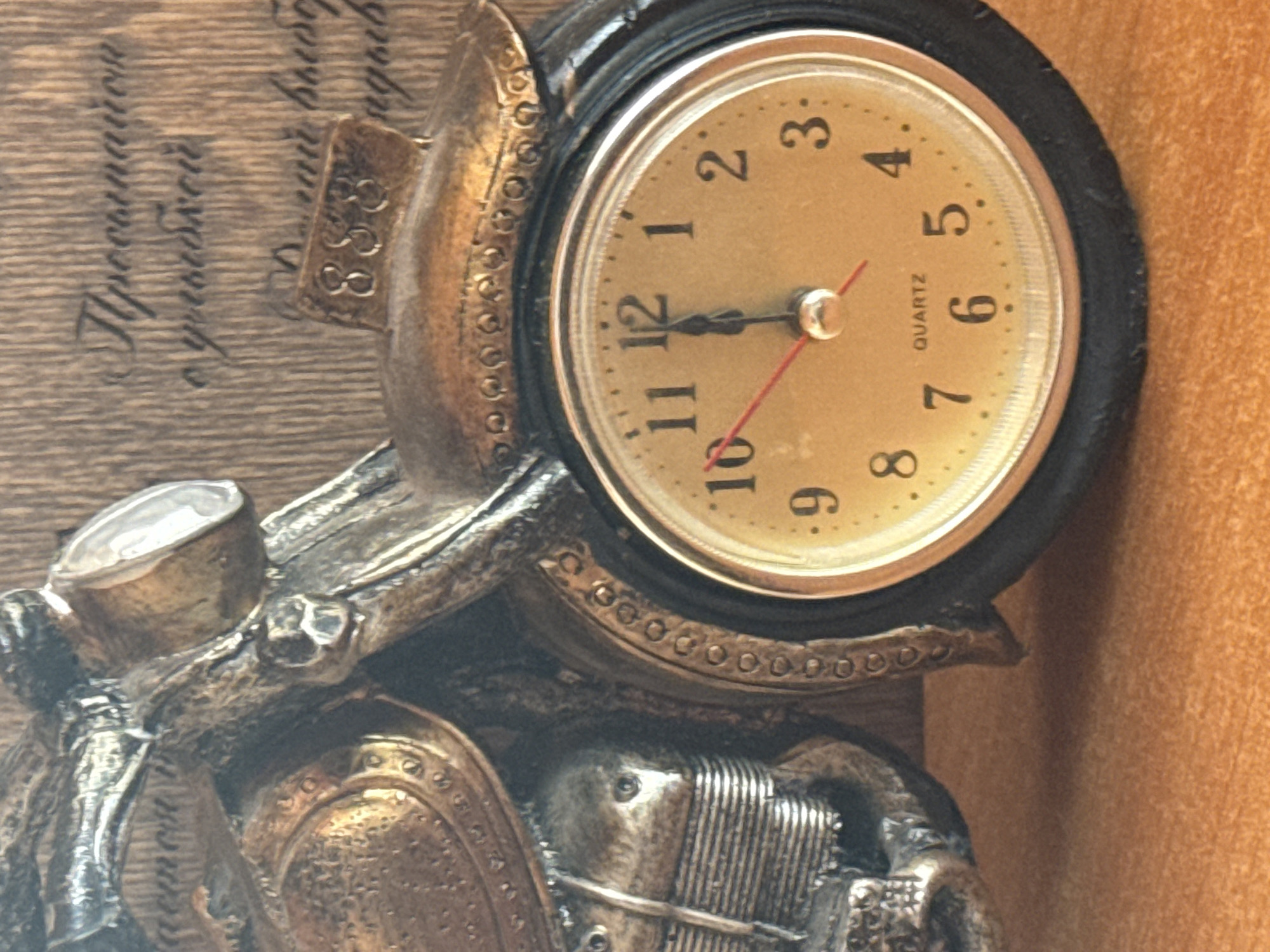 This is a close-up, rotated shot of a decorative clock that appears to be part of a larger sculpture. The clock face is round with a golden dial and black numerals. It shows the time as approximately 10:58. The clock is housed within a dark, possibly metal frame that has decorative embellishments. The clock itself is mounted on a metallic sculpture that resembles a motorcycle, with various mechanical and saddle-like details visible. The background appears to be a wooden surface with some handwritten text in Russian, partially obscured by the sculpture. The overall impression is one of a vintage or steampunk aesthetic. The text on the wooden surface is difficult to fully decipher due to the rotation and partial obstruction, but some legible words include "с улыбкой" (with a smile), and possibly numbers or other inscriptions. The lighting is direct, creating some highlights and shadows on the metallic surfaces. No people are visible in the image.