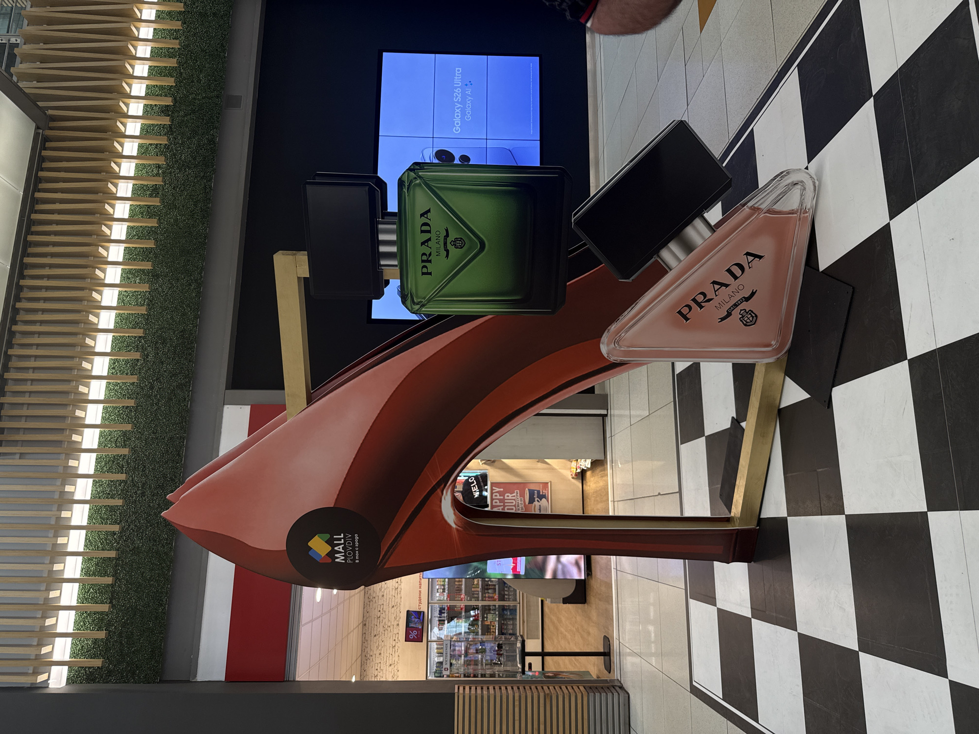 The image captures a promotional display for Prada perfumes, set within the "Mall Plovdiv" shopping center in Plovdiv, Bulgaria. The display features large mock-ups of Prada fragrance bottles atop a stylized, high-heeled shoe structure. The environment includes a tiled floor with a black and white checkered pattern. A shop selling cold beverages with a sign indicating "Happy Hour" is visible in the background. A digital screen displaying marketing content for the Samsung Galaxy S26 Ultra phone is also visible.