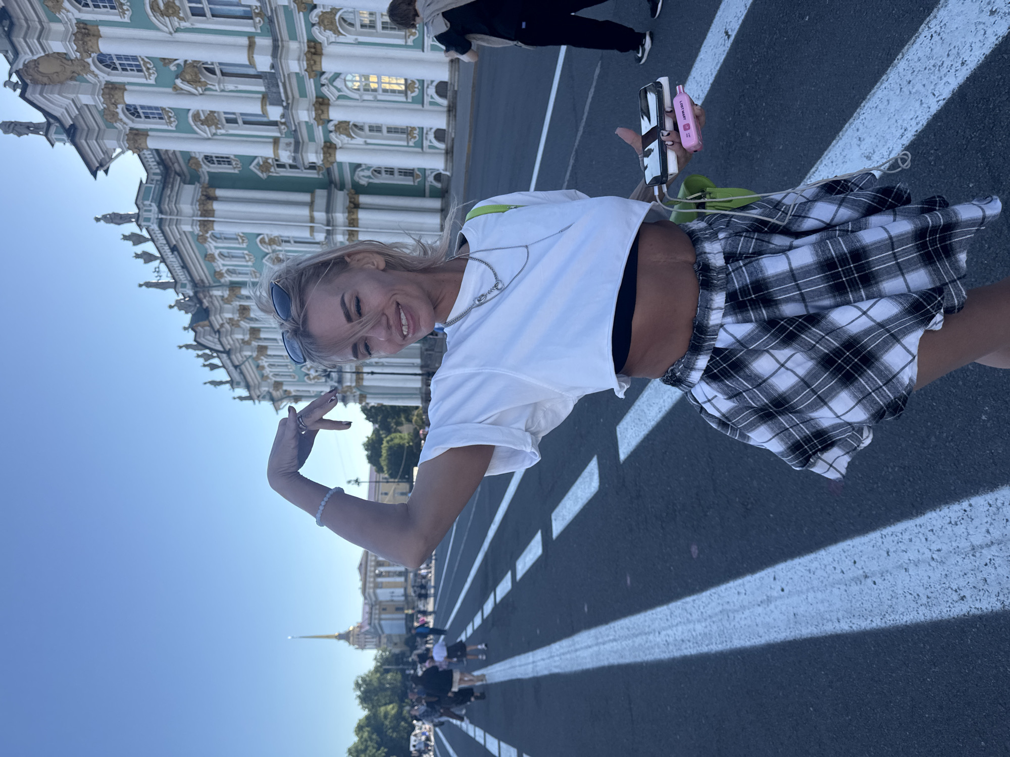 A woman stands on a street in Saint Petersburg, Russia, in front of the Winter Palace. She is wearing a white crop top, a black and white plaid skirt, and sunglasses. She has blonde hair and is smiling. In her right hand, she is holding a phone and a portable charger. The Winter Palace is a large, ornate building with green and white accents. There are other people walking in the background, as well as trees and a clear blue sky. The street is paved with asphalt and has white lane markings.