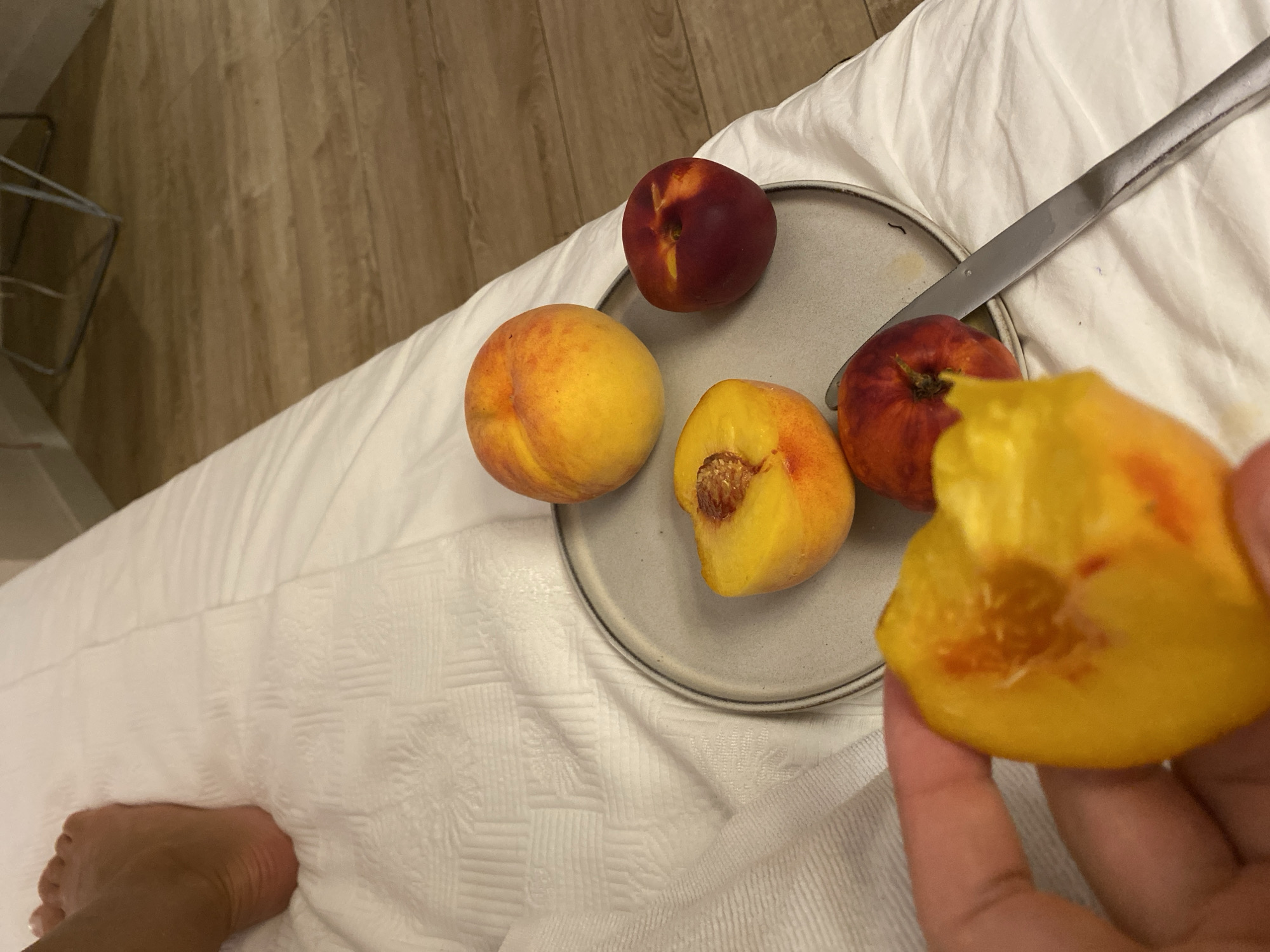A close-up, eye-level shot shows a person's hand holding a partially eaten peach over a plate with several whole and sliced peaches. The peaches are a mix of yellow and red. A knife lies on the plate. The scene appears to be set on a white, textured surface, possibly a bed. In the background, wooden flooring and the leg of a chair are visible. The location is not specified as being in Aksu, Turkey.