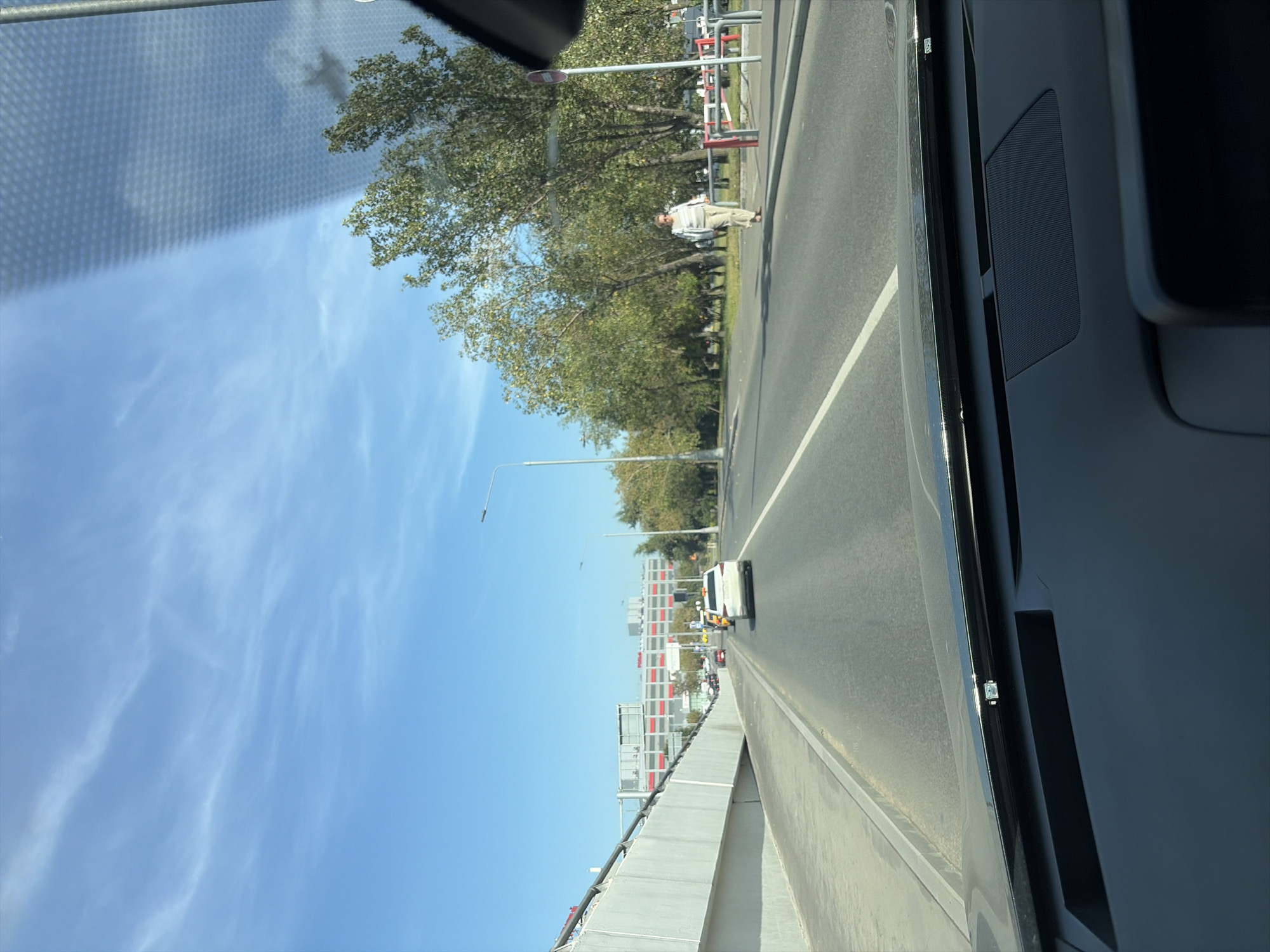 The image shows a view from inside a car on a sunny day, looking out onto a street in Moscow, Russia. The street is paved with asphalt and has white lane markings. On the right side of the street, there is a sidewalk with trees and a pedestrian walking. In the distance, there are buildings and vehicles on the road. The sky is blue with some wispy clouds. The interior of the car is partially visible in the foreground, including the dashboard and windshield.