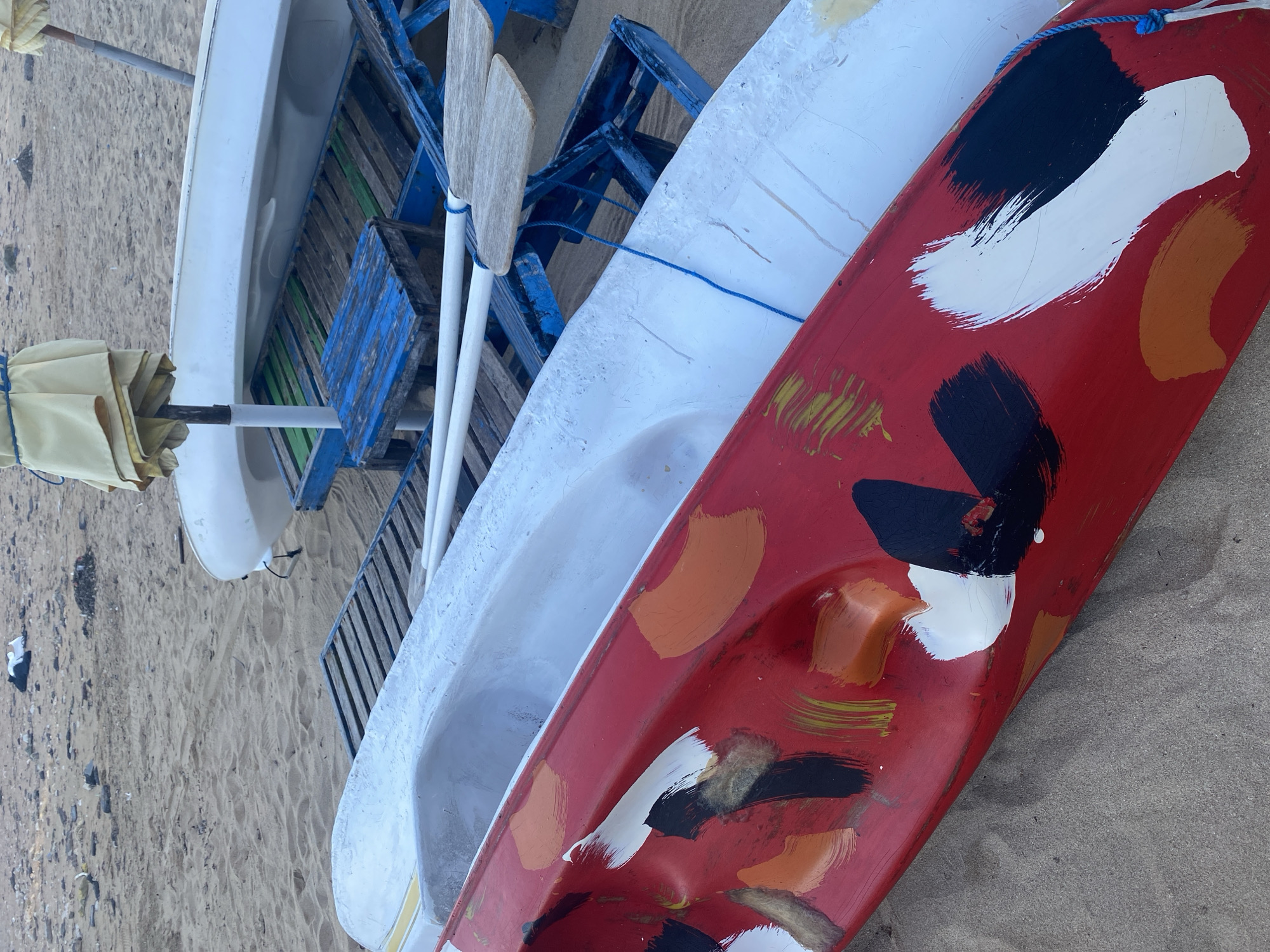 Two kayaks, one red with abstract white, black, and orange paint strokes and one white, are resting on a sandy beach. They are situated on top of blue and green wooden beach chairs or loungers. Partially visible in the background is a white beach umbrella and more beach chairs. The scene is on a beach, likely in Denpasar, Indonesia, indicated by the presence of kayaks and beach furniture typical of coastal tourist areas in the region. No people are visible in the frame.