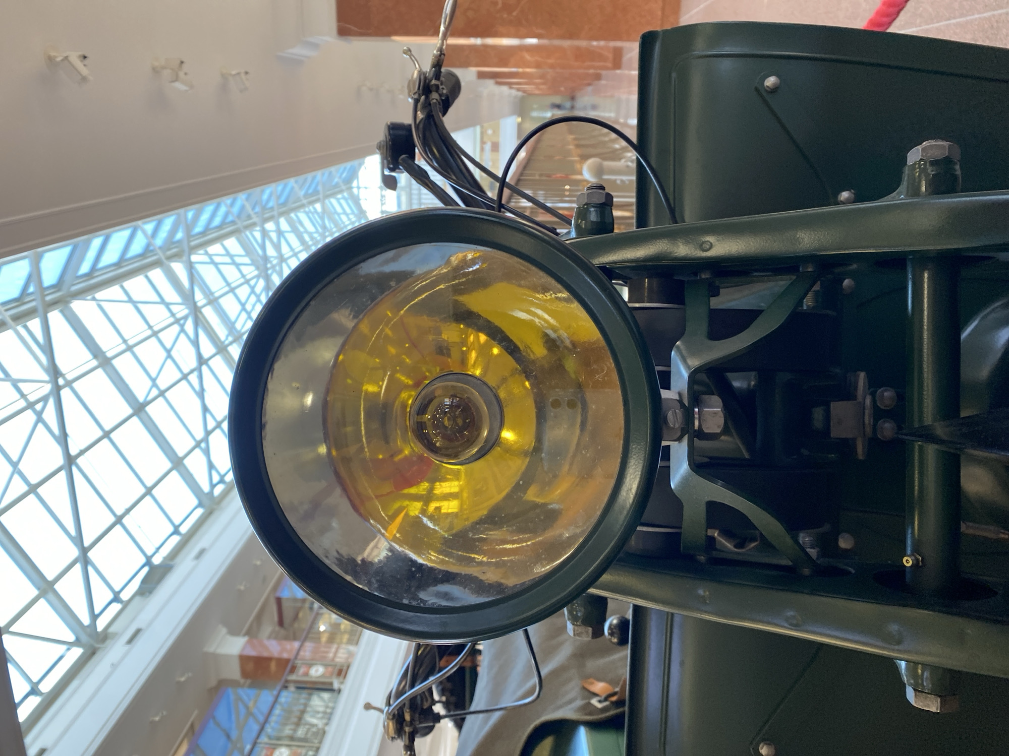 The image is a close-up, eye-level shot of the headlight of a vintage motorcycle, likely on display indoors. The motorcycle is painted a dark green, and the headlight is round with a yellow-tinted lens. The background shows the interior of a building with a glass ceiling, suggesting a museum or exhibition hall. The architecture in the background, with its tall pillars and balconies, is consistent with the Museum of Military Technology in Verkhnyaya Pyshma, Russia. There are no people visible in the frame.