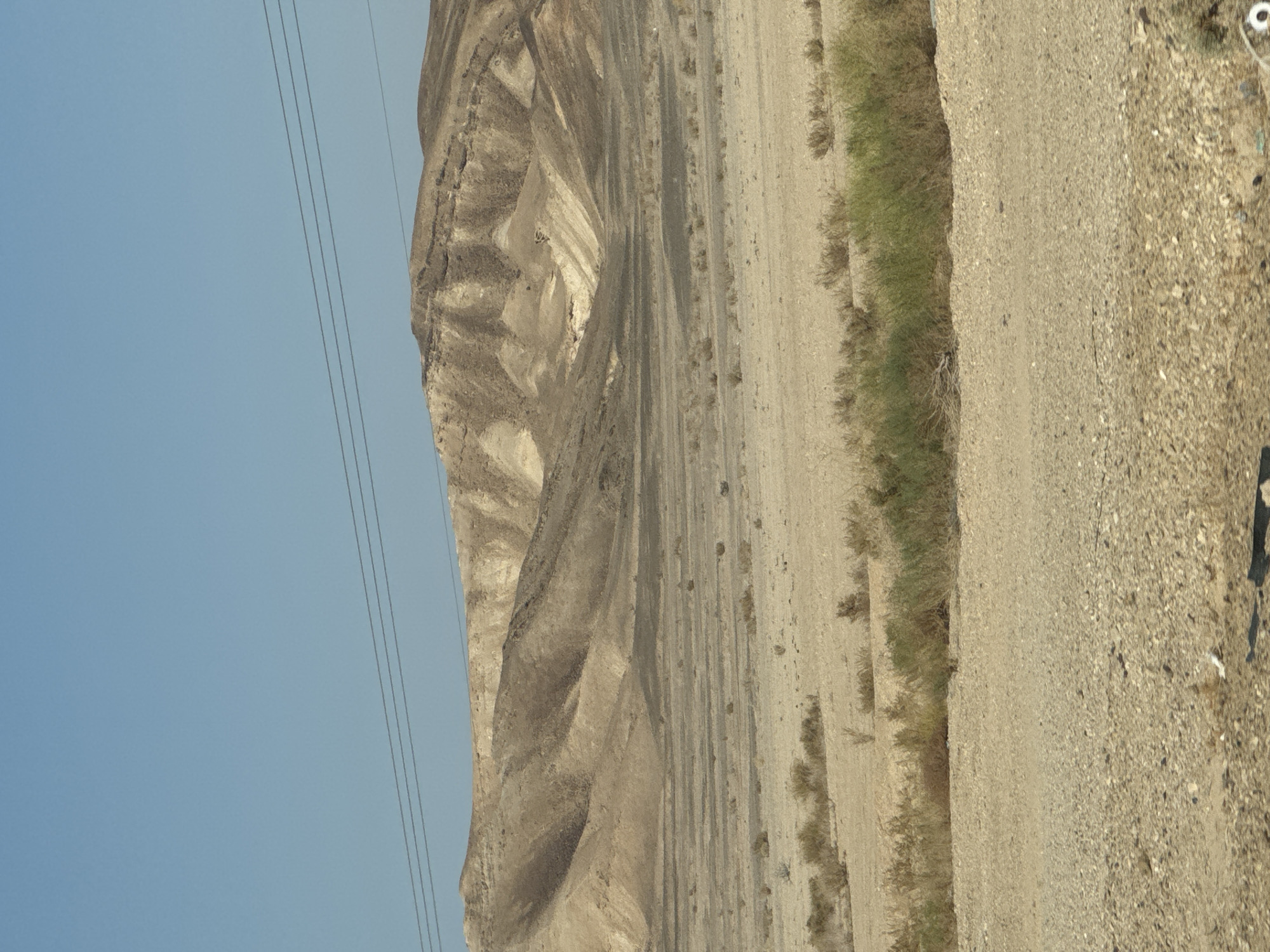 The image depicts a desert landscape under a clear blue sky. In the foreground, the ground is covered with small pebbles and sparse, dry vegetation. Mid-ground shows a wide, flat expanse of desert with scattered dry bushes. In the background, rolling hills with stratified rock formations rise up, displaying shades of tan and light brown. Three dark power lines stretch horizontally across the upper portion of the frame, spanning the distance between unseen poles. The overall scene is arid and uninhabited.