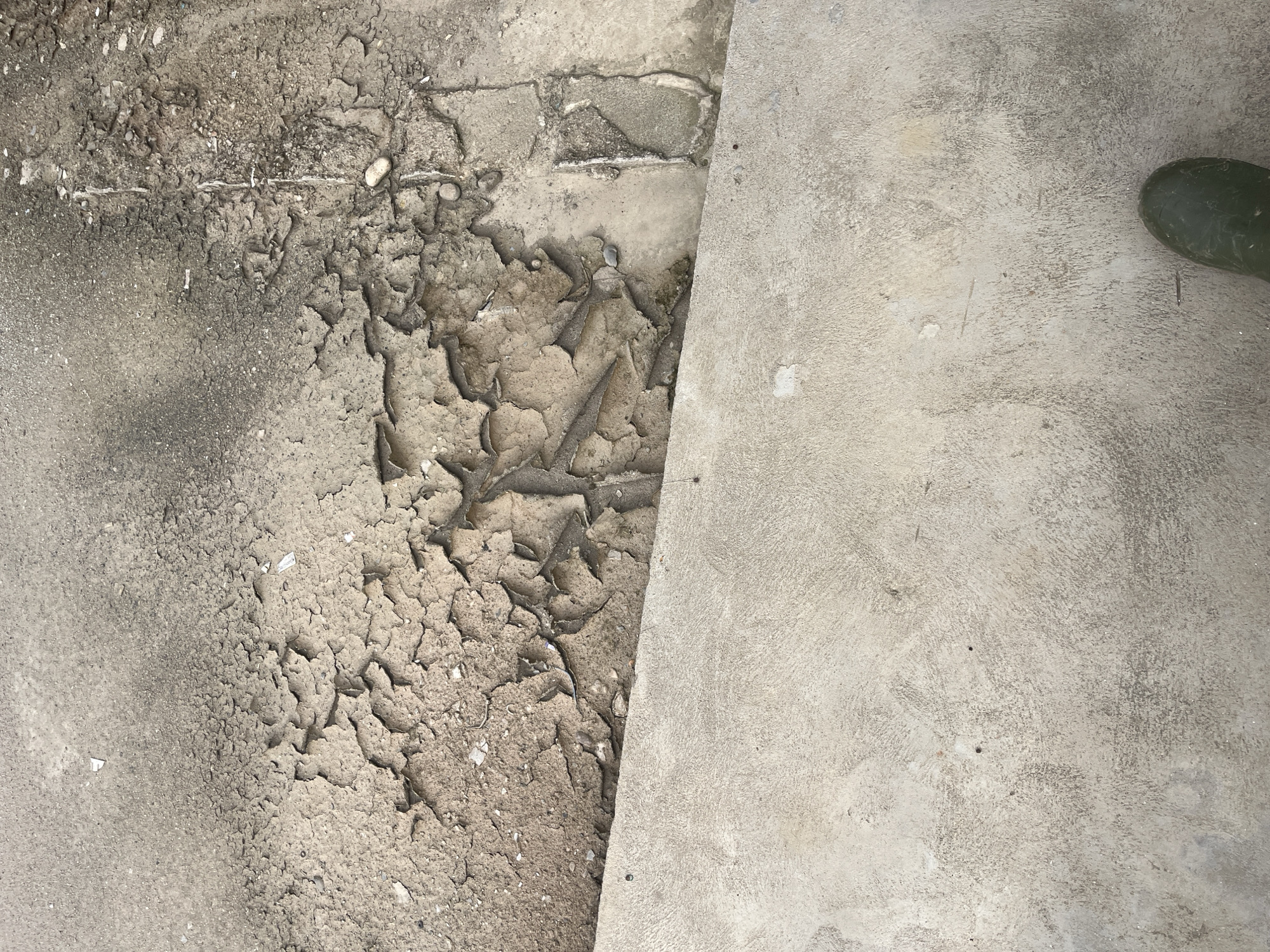 The image shows a close-up, overhead view of a cracked and peeling concrete surface, possibly a sidewalk or pathway, in the city of Buleleng, Indonesia. The texture of the concrete varies, with some areas appearing smooth and the majority exhibiting a rough, weathered finish with prominent cracks and peeling layers. Scattered debris, such as small stones and possibly cigarette butts, are visible on the surface. A distinct line separates this damaged concrete from a smoother, lighter-colored concrete section below, which might be a step or a different section of pavement. In the lower right corner, the toe of a dark green rubber boot is visible, suggesting the presence of at least one person, likely a pedestrian. The lighting suggests an outdoor environment, possibly under daylight conditions.