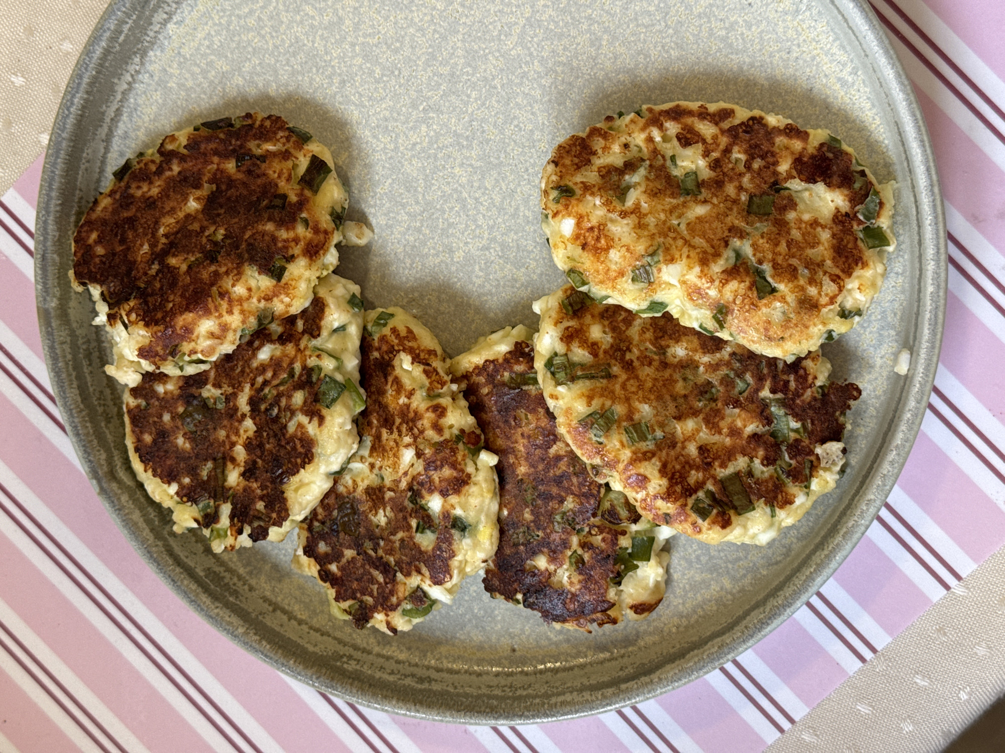 A top-down shot shows several cooked fritters on a gray plate, set on a striped pink and white surface with beige accents. The fritters have a browned exterior and contain green specks, likely herbs.