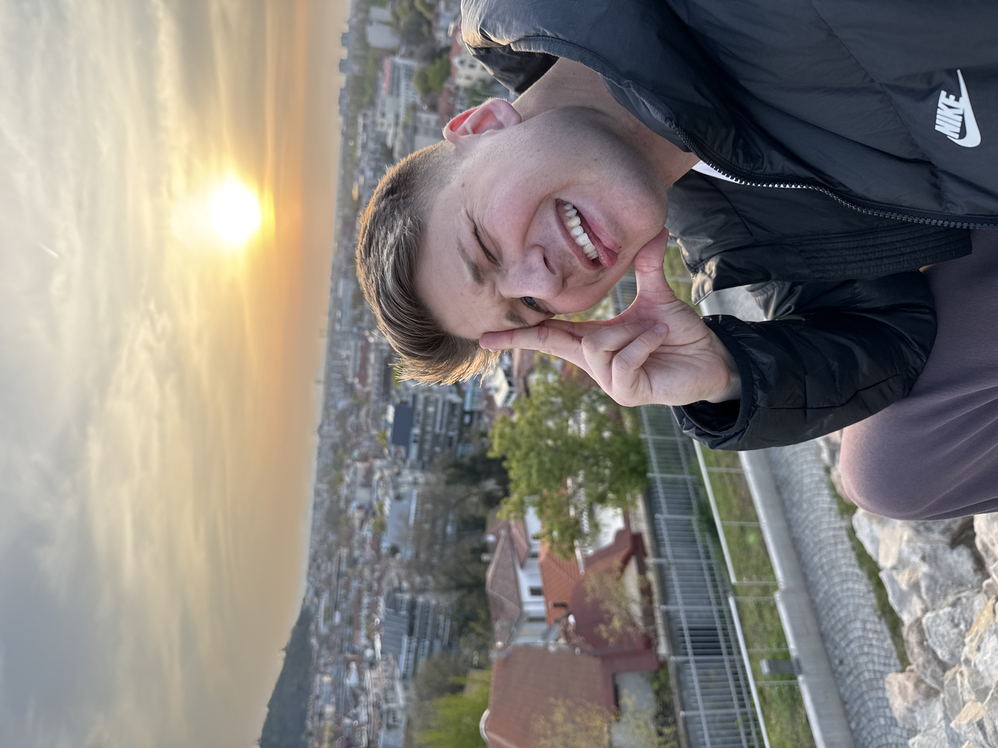 A photo of a person in Plovdiv, Bulgaria.
The person, wearing a black Nike jacket, is smiling with their tongue out and making a 'V' sign with their hand near their face.
The background shows a city with buildings and trees, likely Plovdiv, and a sunset or sunrise. A mountain range is also in the background.
The person is near a stone wall and a metal railing.