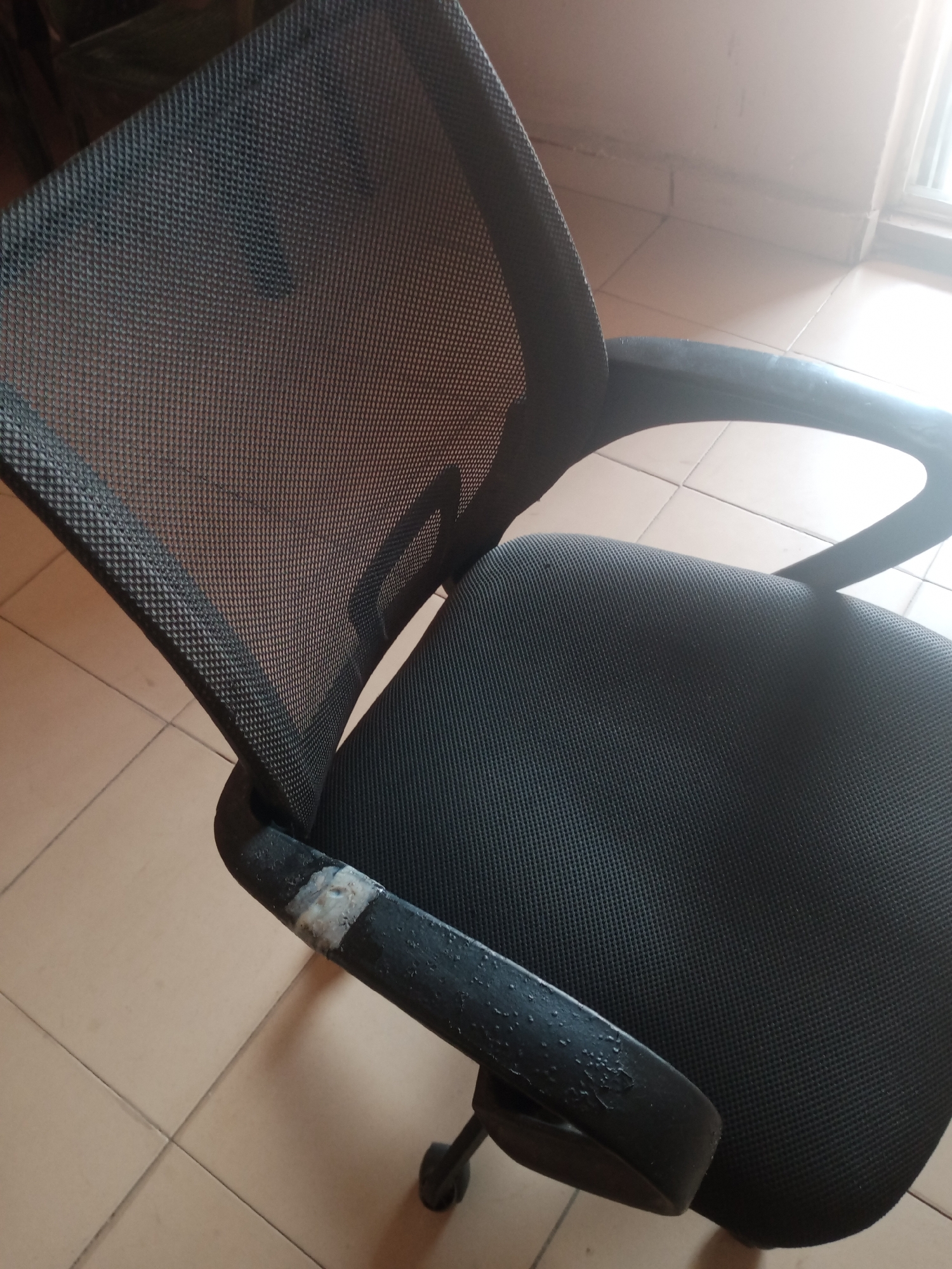 An office chair with a black mesh back and a black fabric seat is depicted. The chair has black armrests, one of which shows signs of wear and tear, with a patch of lighter material exposed and some peeling on the surface. The chair is positioned on a tiled floor that appears to be a light beige or tan color. In the background, a window or doorway lets in bright light, casting a strong glare on the scene. The overall environment suggests an indoor setting, possibly an office or a room in a residential building. The image does not provide any specific information about the city of Jalingo, Nigeria, as it focuses solely on the object and its immediate surroundings. There are no people or actions visible in the image.