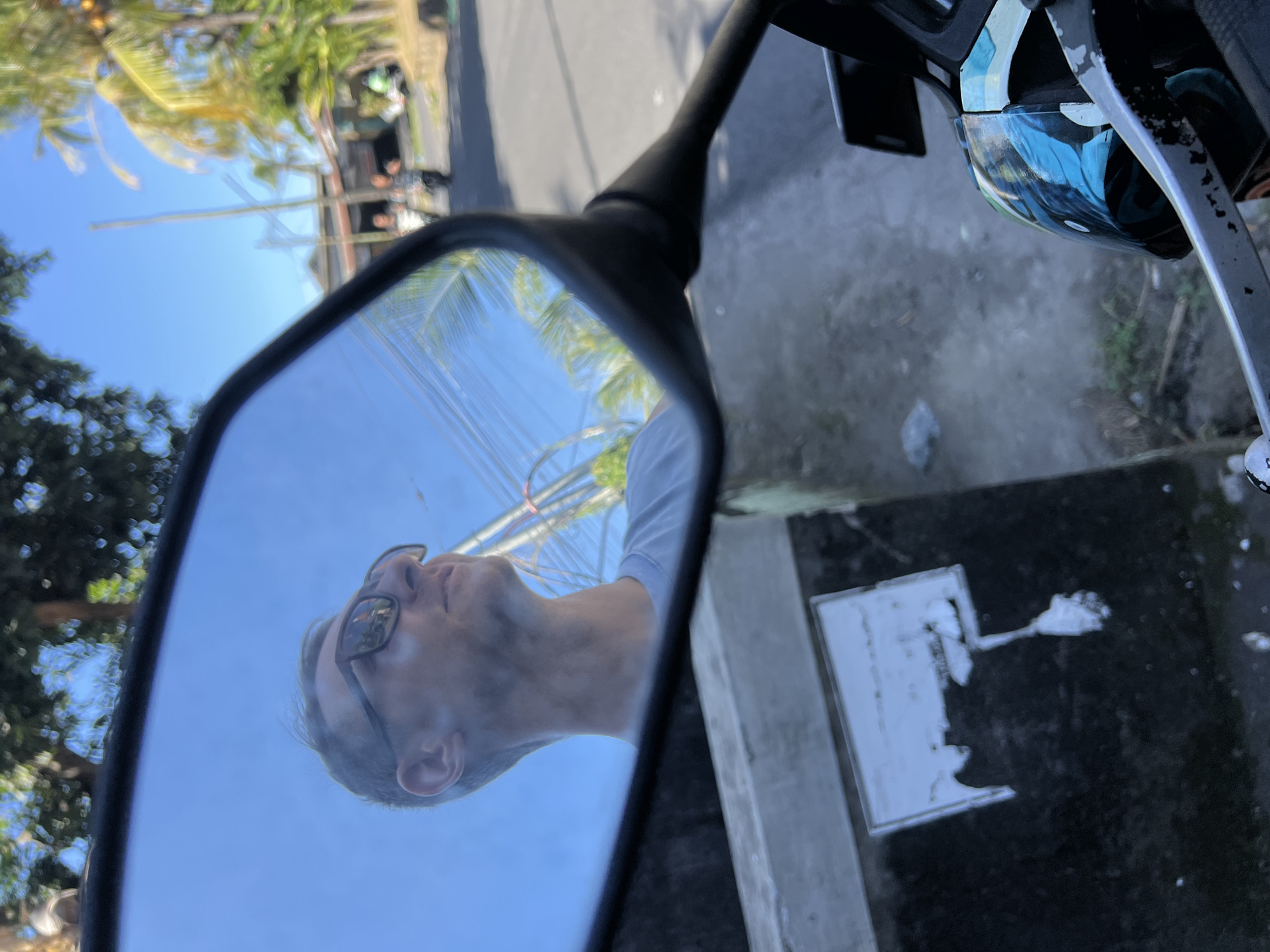 The image shows a reflection in a motorcycle's side mirror. In the reflection, a man wearing sunglasses and a light blue t-shirt looks up and to the right. Behind him, the sky is blue with scattered clouds, and trees with dappled sunlight can be seen. A power line stretches across the background. To the right of the man's reflection, a street scene is visible with palm trees lining the road, a few buildings, and two people walking in the distance. The lower portion of the image shows the motorcycle's handlebar and a decorated helmet. The scene appears to be in Sukawati, Indonesia, given the presence of tropical vegetation and the style of the buildings and power lines.