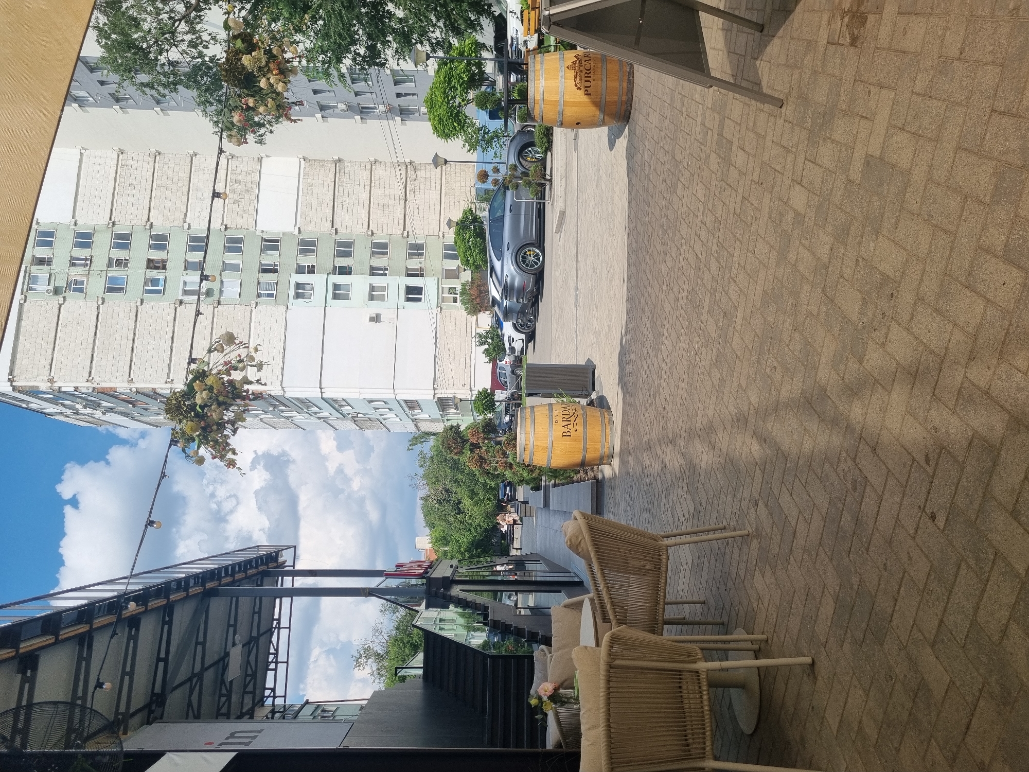 An outdoor seating area with wicker chairs and tables is shown in the foreground of this scene, which is located in an urban environment. Beyond the seating area, a paved walkway leads to a building with a dark exterior and a set of stairs. In the background, a tall apartment building with many windows and a light-colored facade stands adjacent to trees. Several cars are parked on the street, and a wooden barrel sits on the pavement. The sky is visible with scattered clouds.