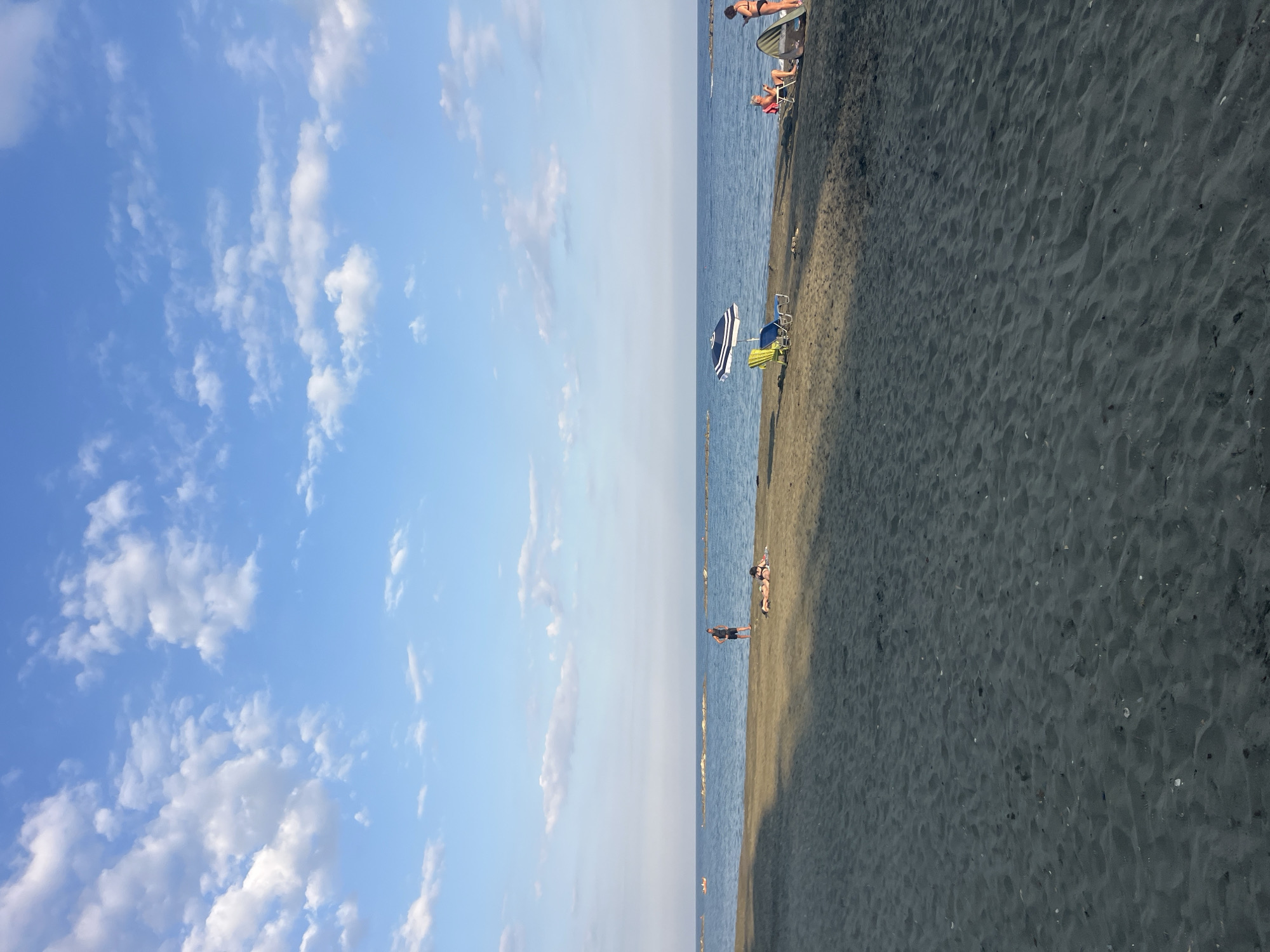 The image depicts a beach scene in Agios Tychona Community, Cyprus. The sand is dark, with a slight sheen, and appears damp in some areas. The sea is a deep blue, with a calm surface and a clear horizon. Several scattered rock formations are visible in the water, extending out from the shoreline. The sky is a bright blue, populated with scattered white clouds.
In the foreground, the dark sand stretches across the frame. A shadow from an unseen object falls diagonally across the sand from the left side of the image.
Further back, on the wet sand near the water's edge, two individuals are lying down. One appears to be a male in dark shorts and a t-shirt, standing and facing away from the camera. Another person, a female, is lying on a towel.
More towards the right side of the image, a blue and white striped umbrella is set up over two beach chairs, one blue and one yellow. A short distance away, a person is seated in a red beach chair, looking down at a device. Next to them, partially obscured by shadow, is a green windbreak or tent. A person in a dark swimsuit is standing nearby, looking out towards the sea.
In the distance, two small boats are visible on the water, and a few more rock formations are spread along the horizon. The lighting suggests it is late afternoon or early evening, with the sun casting long shadows.