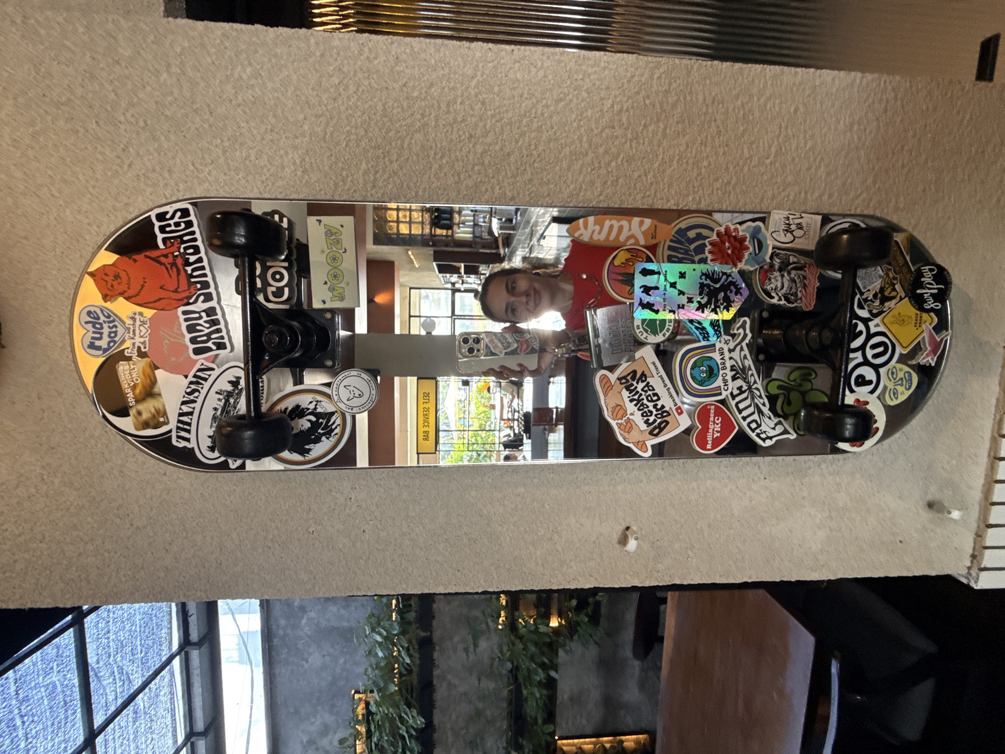 The image shows a reflective skateboard mounted on a textured wall. The skateboard is covered with various stickers. A young woman is reflected in the skateboard's surface, taking a picture of it with her smartphone. The background reflection shows an interior with a window and plants. Based on the stickers and the general aesthetic, the location is Uluwatu, Indonesia.