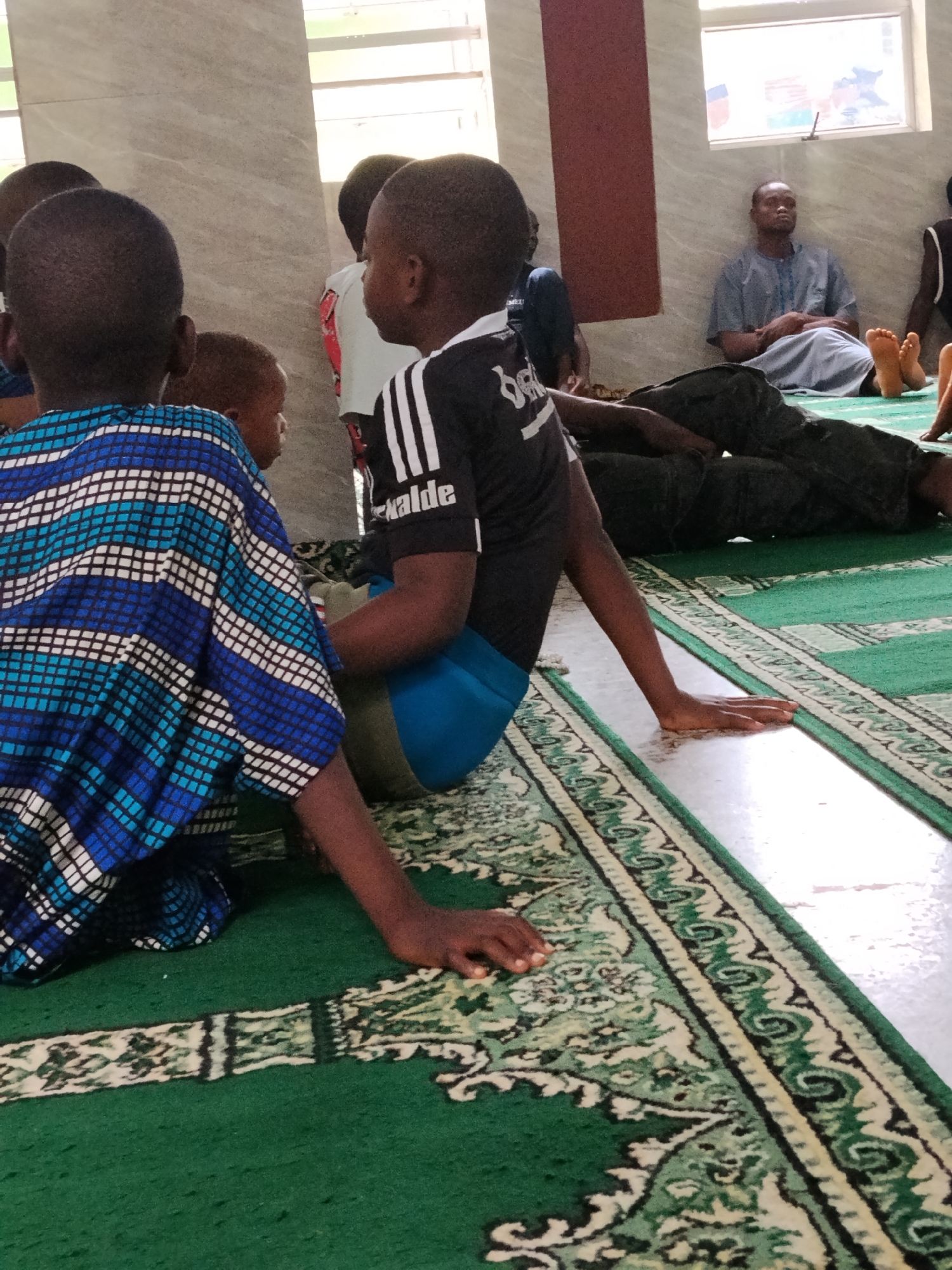 The image depicts an indoor scene with several people, primarily children, seated on a green patterned carpet. The setting appears to be a mosque or a religious educational space, indicated by the prayer mat-like flooring and the communal gathering. Natural light streams in from windows visible on the light-colored walls, suggesting it is daytime.
In the foreground, a child wearing a distinct blue, white, and black patterned garment is seen from behind. To their right, another child, viewed in profile, wears a black sports t-shirt with white stripes on the sleeve. On this shirt, the words "Schalke" and "kalde" (partially obscured) are visible. Both children are seated on the carpet, with their hands resting on the floor.
Further in the background, an adult male is seated against a light-colored wall with a brown vertical accent, looking forward. He is dressed in a light blue traditional tunic. Other individuals are also present, some lying down or seated on the carpet. The overall activity seems to be a quiet gathering, possibly for a lesson, prayer, or rest. The atmosphere is calm, and the individuals appear attentive or relaxed. The perspective is low, focusing on the children and the intricate patterns of the carpet.