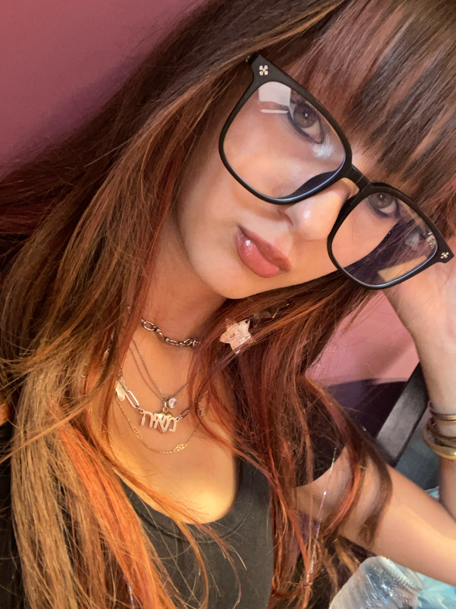A woman is pictured from the chest up, wearing black-rimmed glasses and multiple necklaces. Her hair is long and has streaks of red and blonde. The background is a solid pink color. The image was taken indoors. The city of Raanana, Israel is not depicted in this image.