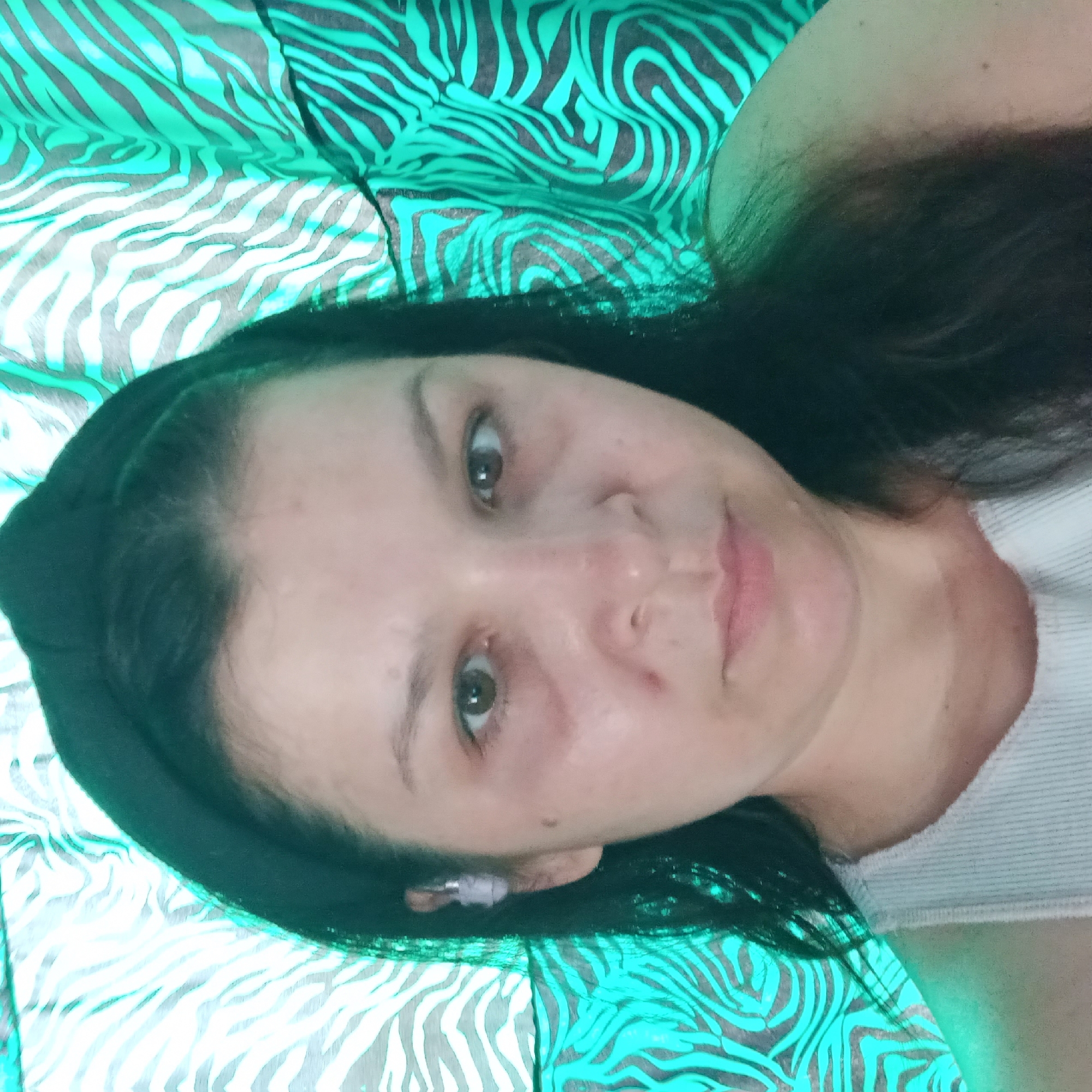 A head and shoulders selfie shows a woman with long, dark hair and a black headband. She is wearing a light-colored ribbed tank top. The background is a green and black zebra-print fabric, which appears to be a curtain or drapery. The lighting is uneven, with a strong green cast, particularly on the left side of the image. The woman's skin shows some redness and puffiness under her eyes. She is looking directly at the camera. There is no indication of the city of Klyntsi, Ukraine in the image.