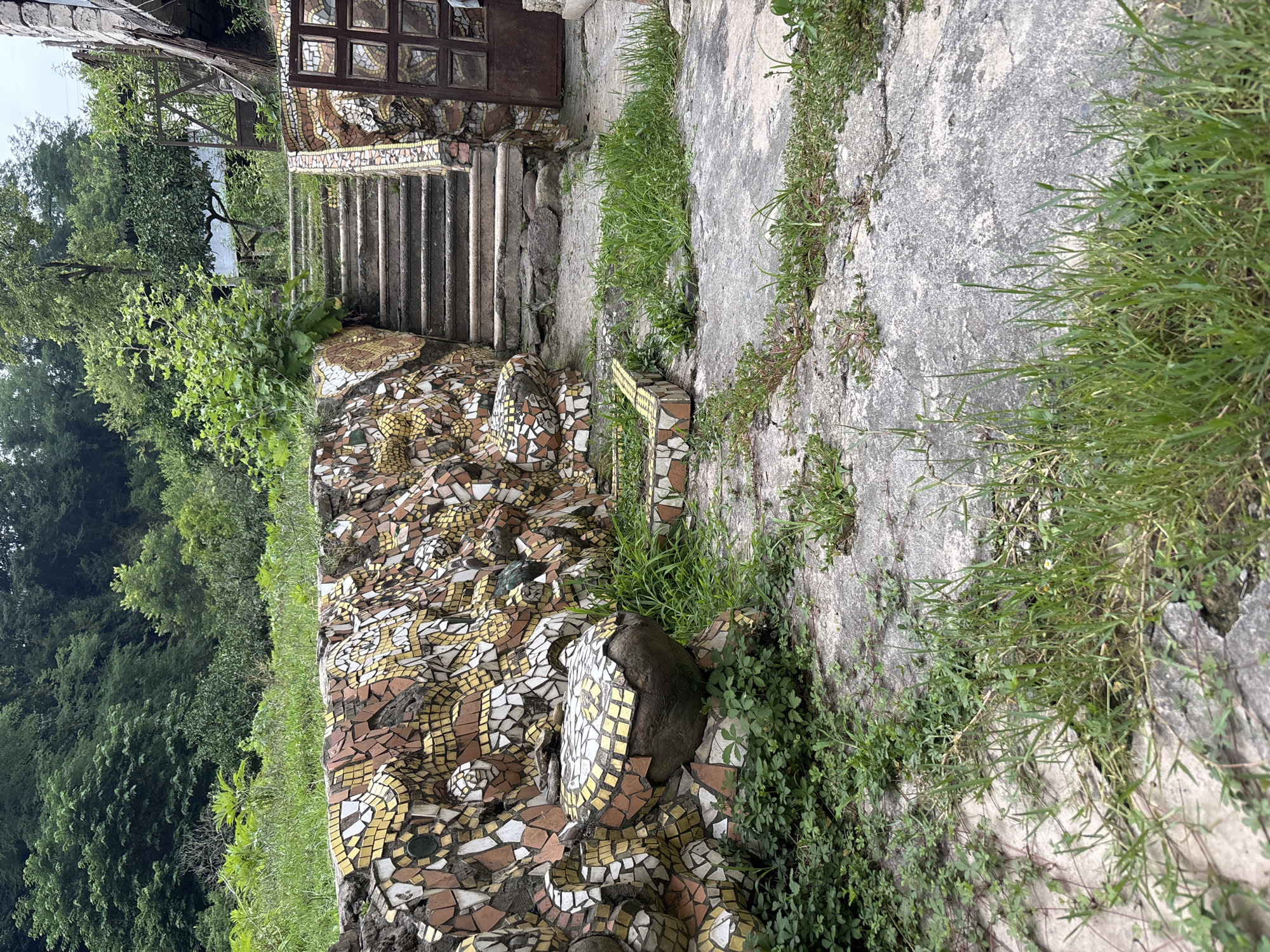 An outdoor stone staircase ascends a gentle slope. The staircase is flanked by a rough, uneven wall decorated with colorful mosaic tiles in shades of yellow, white, and brown. The wall has bulbous, sculpted sections resembling animal forms, also covered in mosaic. Overgrown green grass and weeds sprout from cracks in the concrete pathway leading to the stairs and around the base of the wall. In the background, lush green trees and foliage form a dense forest. A rusty metal gate is visible to the right of the staircase, partially obscured by the wall and vegetation. The scene appears to be in a state of disrepair, with the mosaic art being the most prominent and well-maintained feature.