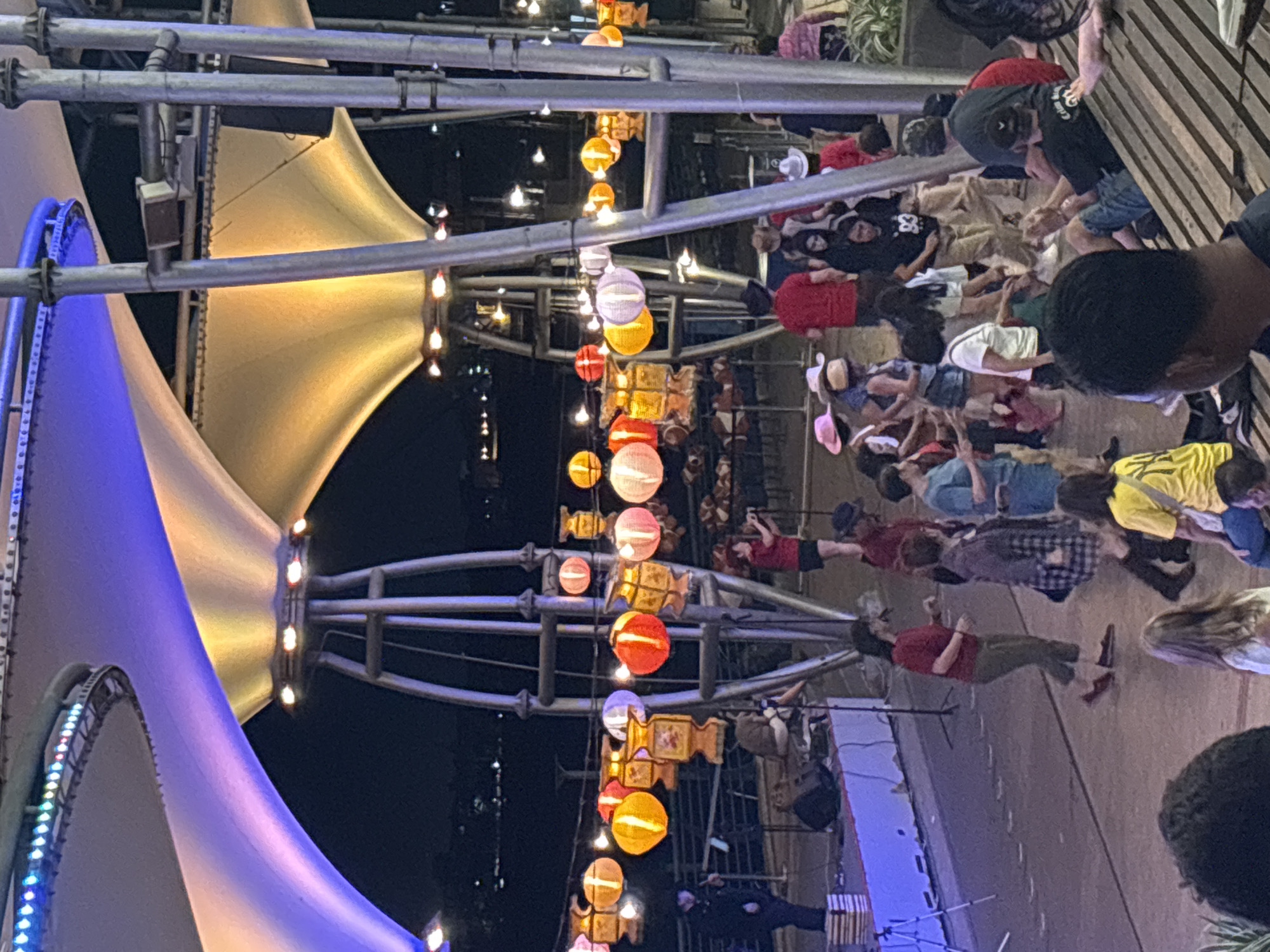 The image depicts a nighttime scene in North Jakarta, Indonesia. Strings of colorful lanterns hang overhead, illuminating an outdoor gathering of people on what appears to be a pedestrian walkway. The lanterns are round and feature various colors and designs, including cylindrical shapes with decorative elements. The walkway is lined with low concrete barriers and metal railings. A large tensile structure, partially illuminated with blue and yellow lights, is visible on the left side of the frame. The crowd consists of men and women of various ages and clothing styles. Some appear to be seated or lying on the ground, while others are standing. The environment indicates an event or festival taking place in a public space.