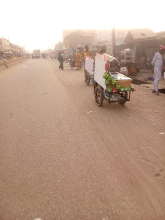 A dusty street scene features a mobile vendor cart laden with snacks. The cart, painted white, has two large wheels and is being pushed along a paved road. Other pedestrians and vendors are visible in the background, adding to the bustling street atmosphere. Buildings line the road, creating a sense of urban density and activity. The lighting suggests a hazy or overcast day, with a yellowish hue permeating the scene.