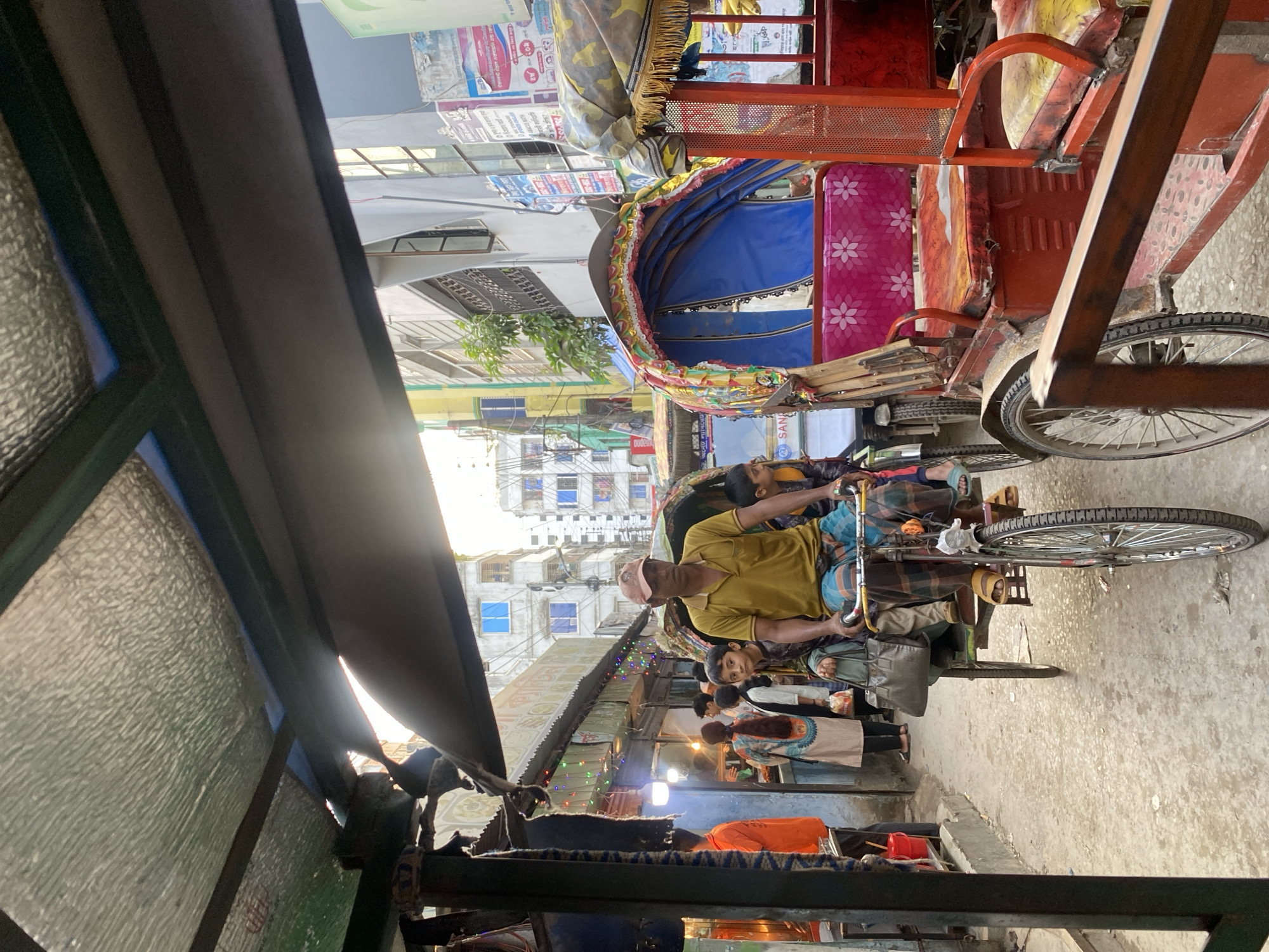 The image shows a street scene in Dhaka, Bangladesh, viewed from inside a rickshaw. A rickshaw driver wearing a yellow shirt and a cap is pulling his vehicle in a narrow street. The rickshaw has a canopy with colorful decorations and a pink-flowered seat. Beside the rickshaw, pedestrians are walking on the pavement. Buildings line the street, with storefronts illuminated by colorful lights. The foreground shows the structure of the rickshaw in which the picture is being taken.