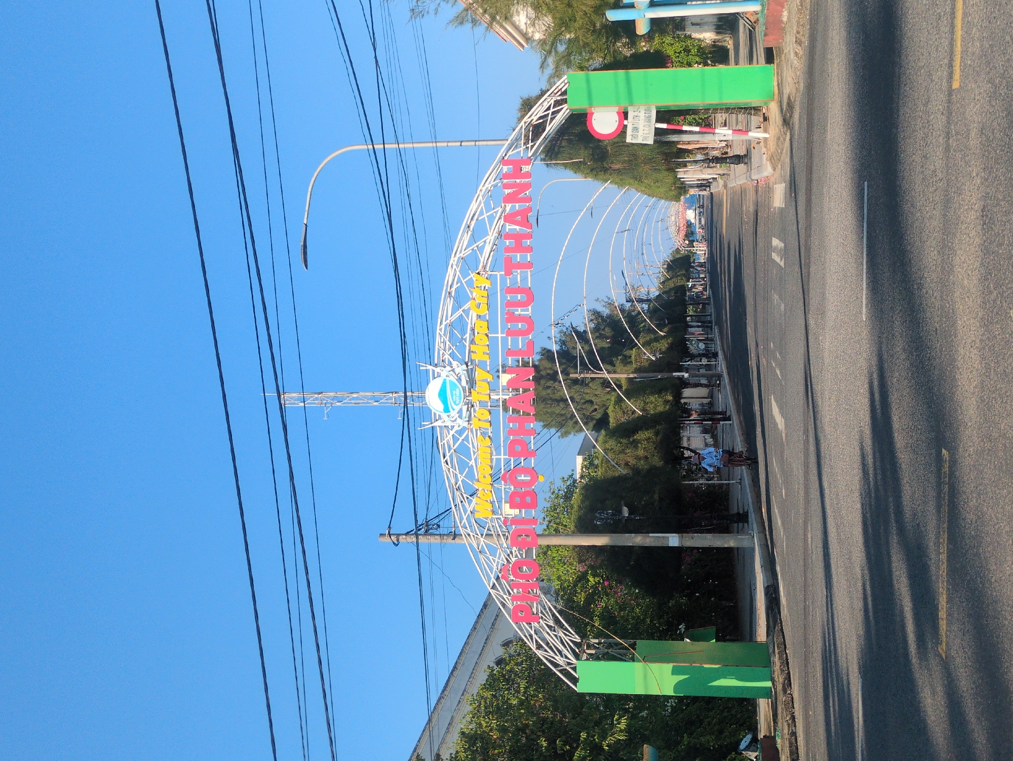 An eye-level outdoor shot depicts an archway over a road, with text in Vietnamese, "Welcome To Tuy Hòa City ĐẢO BẠCH TUYẾT THANH". The archway is constructed of white lattice with red and yellow lettering, with a blue and white emblem.
The scene is located on a street in Tuy Hòa, Vietnam, with the perspective looking down the road. The road is asphalt, with a yellow line on the right and painted white lane markers.
Power lines are visible in the upper left against a clear blue sky. Street lamps stand to the side. Green rectangular structures hold up the arch. Trees and foliage are visible on either side of the road beyond the arch. A motorcyclist is on the left side of the road.
