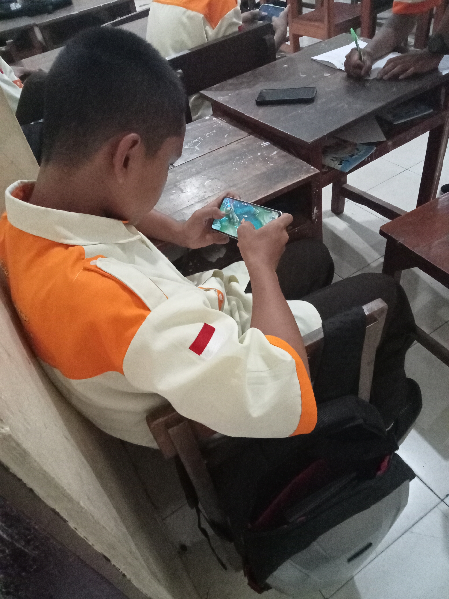 In Secang, Indonesia, a young male, seen from the shoulder up, is engrossed in playing a game on his smartphone. He is seated at a wooden desk, wearing a collared shirt with orange and white panels, and a small Indonesian flag patch on the sleeve. His hands grip the phone, displaying a colorful game interface. Another person's hand, holding a green pen, is visible in the background, writing on a piece of paper on a similar desk. The setting appears to be a classroom or study area, with additional wooden desks and chairs present. The floor is tiled. A black backpack is situated on the floor near the young male.