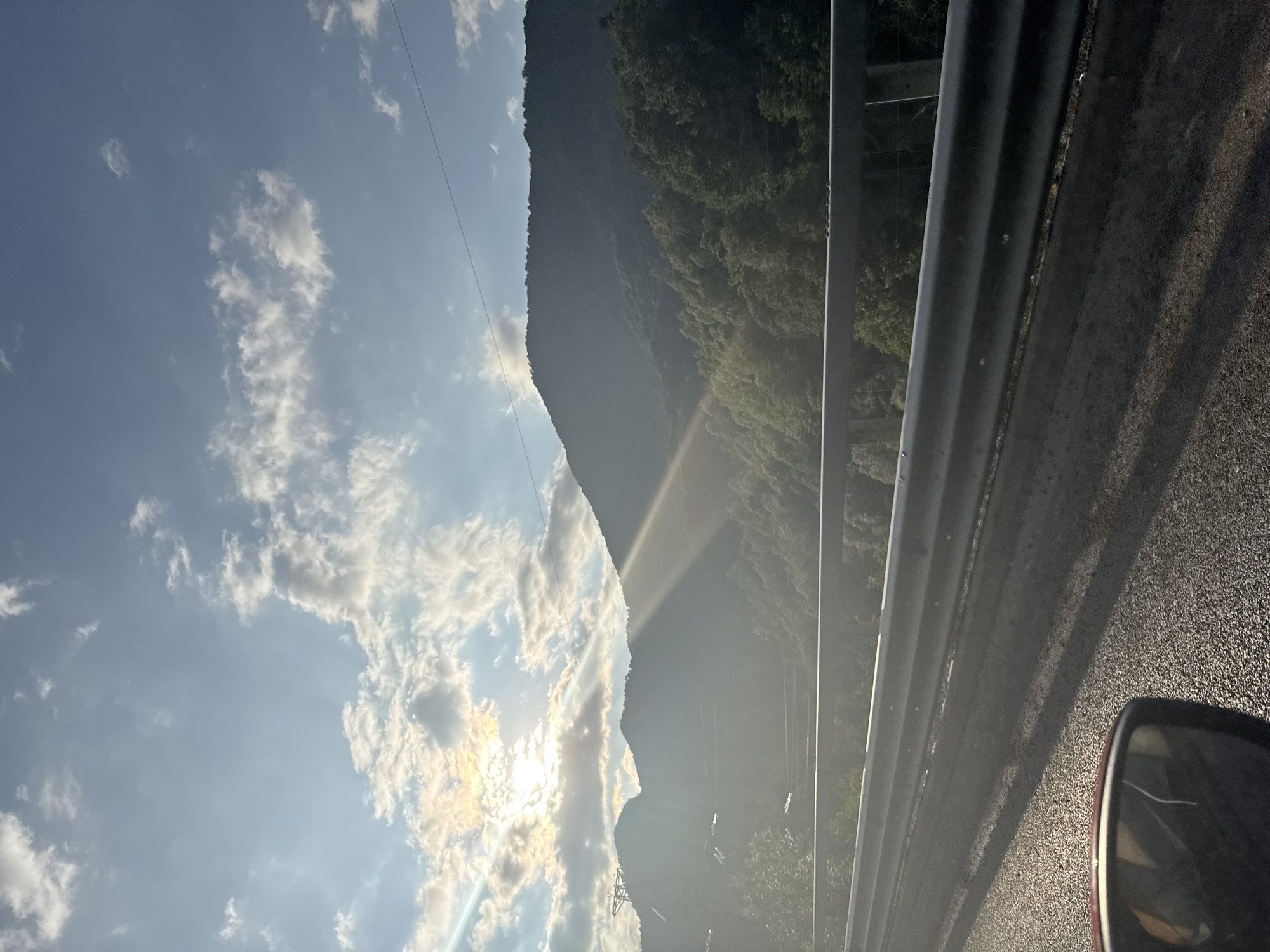 The image depicts a scenic view from a vehicle, likely in Krasnaya Polyana, Russia. The foreground shows the asphalt road and a silver guardrail, with a sliver of a car's side mirror visible on the right. Beyond the guardrail, a steep, tree-covered mountain slope rises dramatically, casting a shadow. The sky is partly cloudy with the sun shining through, creating a bright, ethereal light that beams down through the clouds and onto the mountain. The time of day appears to be late afternoon or early evening, as suggested by the low angle of the sun and the long shadows. The overall mood is one of natural beauty and tranquility, with a sense of vastness conveyed by the towering mountains and expansive sky. The lush green of the trees suggests a temperate season, possibly spring or summer. No text is visible in the image.