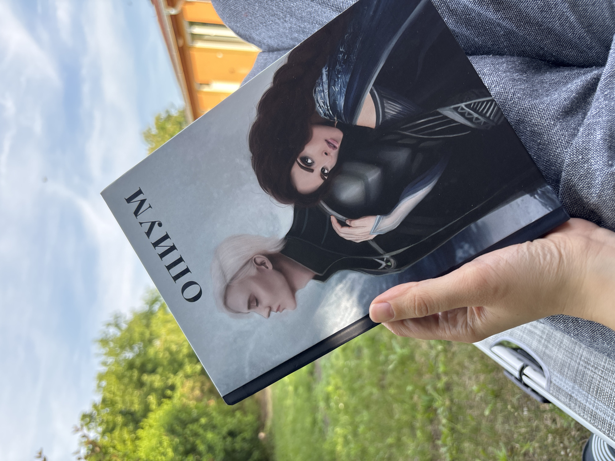 A vertical medium shot shows a person holding a book with a cover featuring two illustrated characters. The character on the left has long dark hair and is looking towards the viewer with large, expressive eyes. She is wearing dark armor with blue accents. The character on the right has short, light-colored hair and is looking down with a profile view. This character is also wearing dark attire. The cover is mostly dark, with a gray background behind the characters. To the left of the characters, in large black serif font, is the word "МАЛИПНО". The background of the photograph shows a blurred view of green foliage and a bright blue sky with wispy white clouds. In the upper left corner, out of focus, is a building with an orange roof. The lighting suggests it is daytime, likely sunny. The book is held in the foreground by a person's hand and arm, which are partially visible on the right side of the frame. The person is wearing a textured gray garment. The overall scene appears to be outdoors in a natural setting, possibly a park or garden.