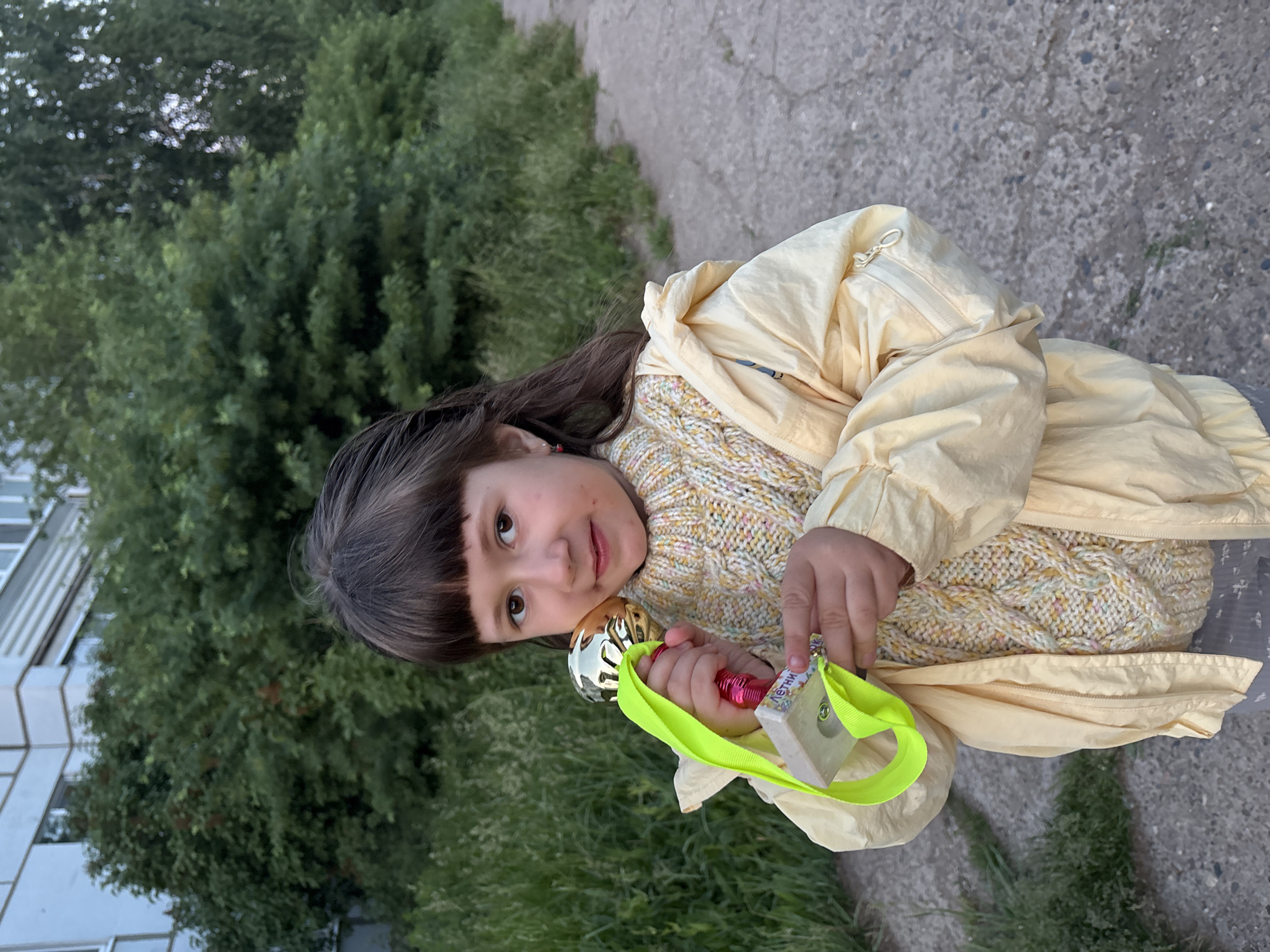 A young girl stands outdoors in Chaikovsky, Russia, holding a toy microphone and a small, rectangular object attached to a neon green strap. She is wearing a yellow jacket over a multicolored sweater and has dark brown hair with blunt bangs. The background includes green bushes and trees, as well as a gray sidewalk and part of a building.