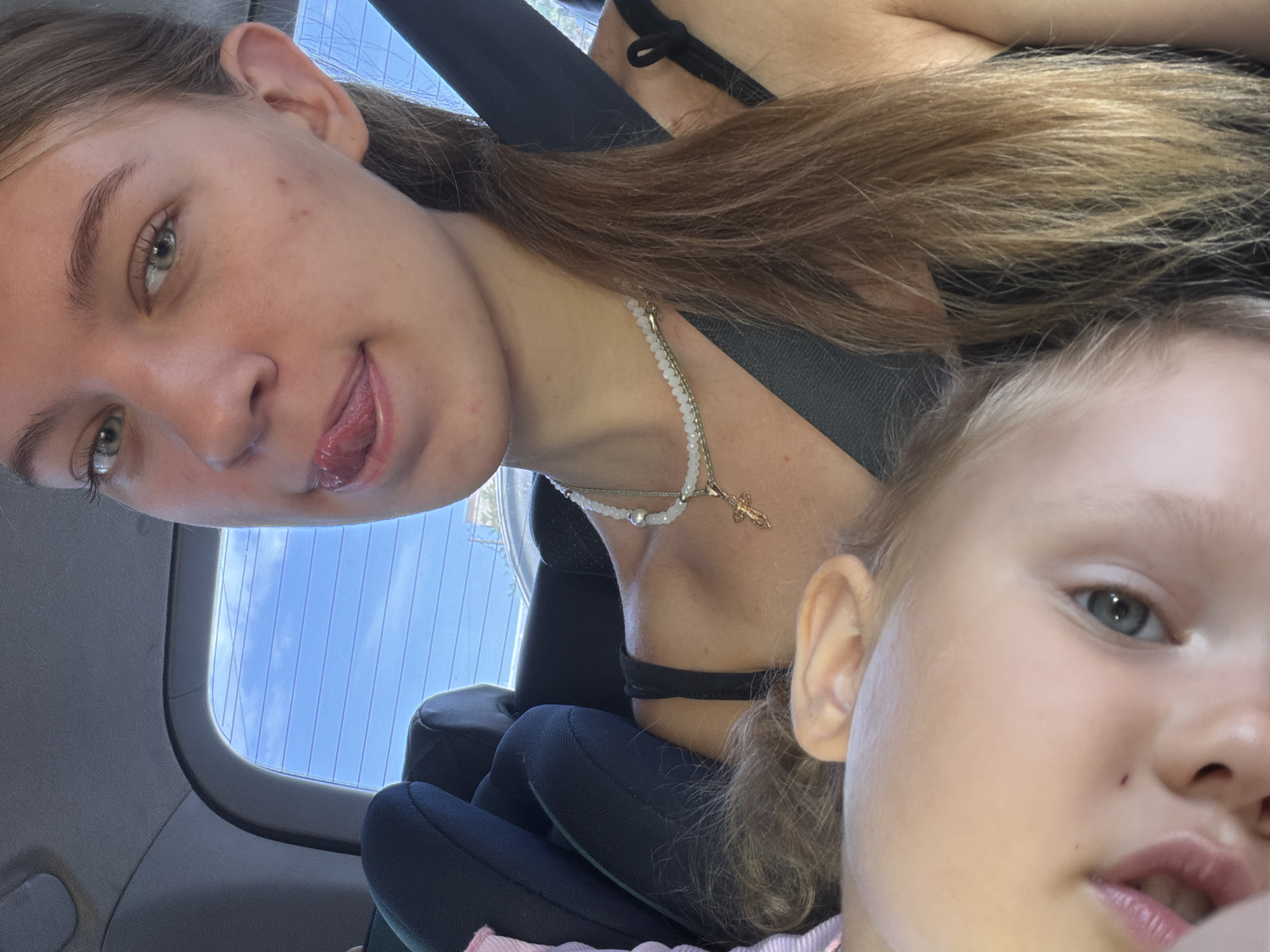 In Rostov-on-Don, Russia, two individuals are seated in the back of a vehicle. The primary subject, positioned in the foreground and to the right, is a young woman with light brown hair, wearing a black top and a necklace with a cross pendant. She is looking towards the camera with her tongue sticking out playfully. Partially visible in the lower left corner is a baby, whose head is tilted, with one eye and part of their mouth visible. The background shows a car interior with a child car seat and a window displaying a bright blue sky with some clouds, suggesting daytime. The scene appears casual and is likely taken from inside the vehicle.
