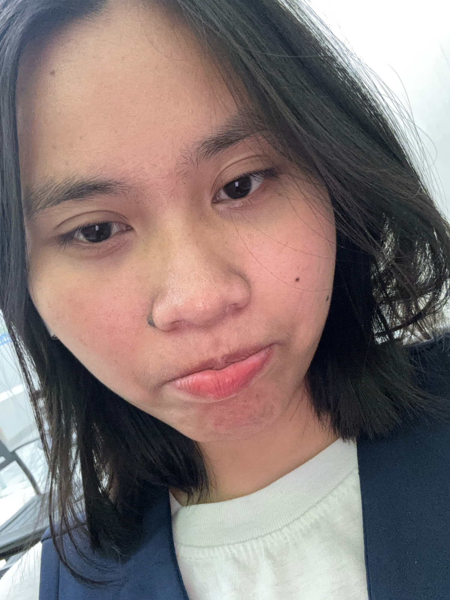 The image is a close-up, selfie-style photograph of a young person's face, captured from a slightly downward angle. The subject has dark hair, parted in the middle and falling around their shoulders, with some strands framing their face. Their skin appears to have a slight redness or blush, particularly on the cheeks. They have dark eyebrows and dark eyes. A small black speck, possibly a beauty mark or blemish, is visible on their right cheek, and another is present on their upper lip. Their lips are slightly parted and appear to have a pinkish hue. The person is wearing a white t-shirt under a dark blue vest or jacket. The background is out of focus and appears to be an indoor setting with some diffused light, suggesting it could be a public space or an office, possibly in East Jakarta, Indonesia, given the inferred location. There are no other individuals or prominent objects in the immediate foreground or background, focusing the attention entirely on the subject's facial expression, which is neutral to slightly pensive.