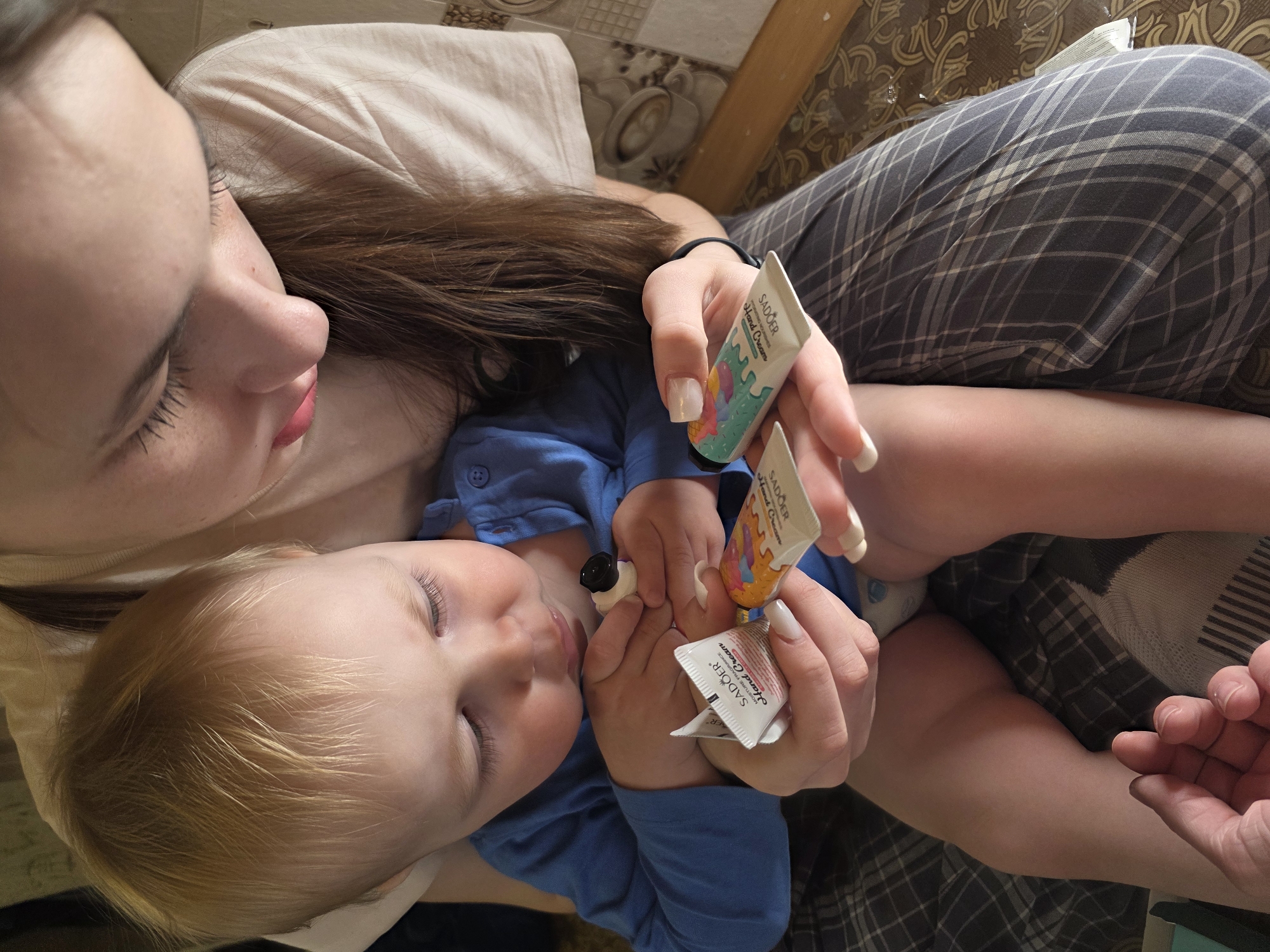 A woman lies on her back on a bed in an indoor location in Chaikovsky, Russia. She has long brown hair and is wearing a light-colored shirt and gray plaid pants. A young child with blond hair lies on her chest. The woman holds two tubes and a foil packet of "SADOER Hand Cream." A second adult hand is visible at the bottom right corner of the image. The background includes a light-colored pillow and a brown patterned surface.