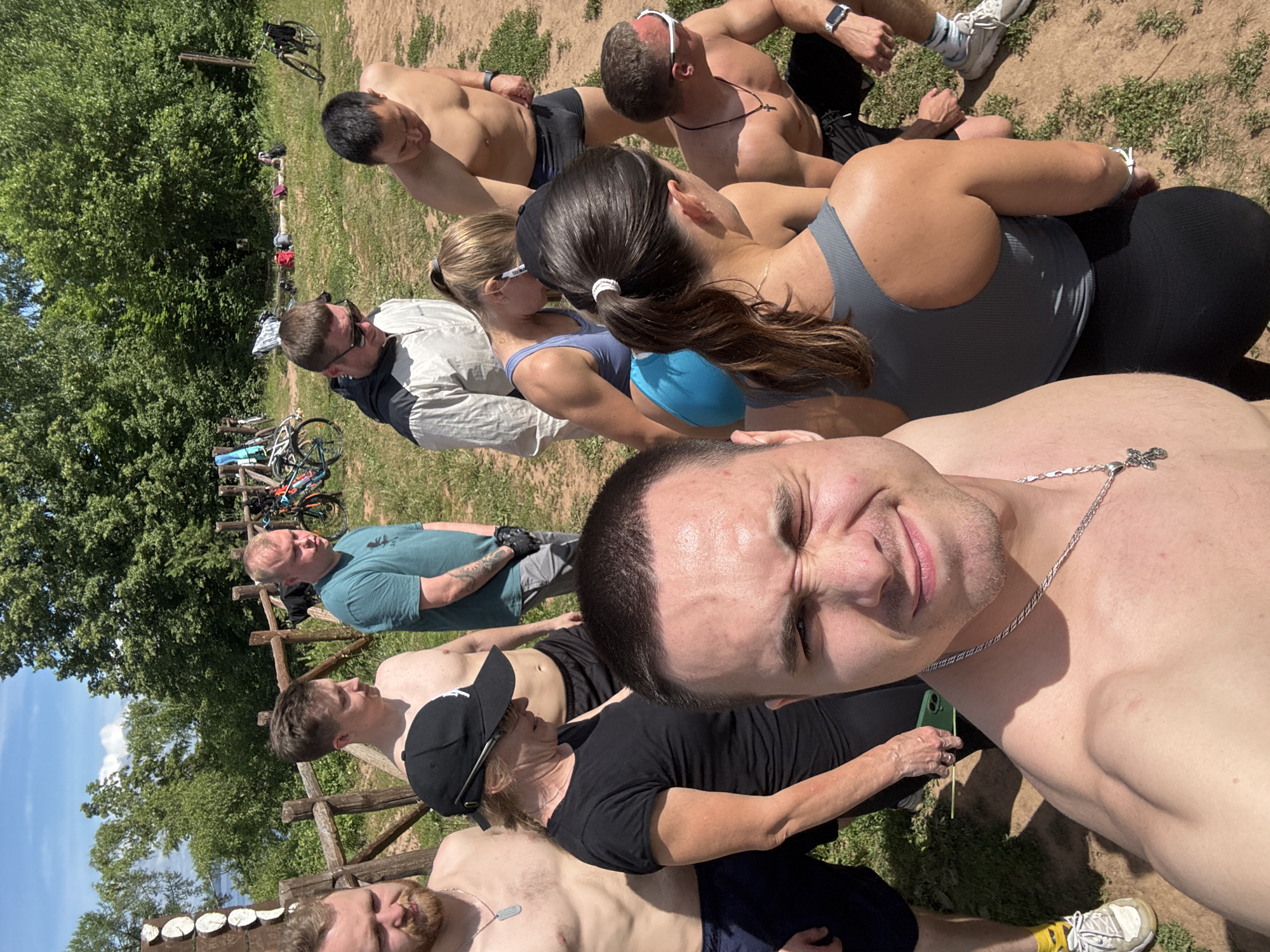 A group of eight individuals, predominantly shirtless and casually dressed, are gathered outdoors on a sunny day in what appears to be a rural or park-like setting. The foreground is dominated by a selfie taken by one of the men, who is looking directly at the camera with a winking expression. He is wearing a silver chain with a cross pendant. To his left, a woman in a black t-shirt and a black baseball cap is holding a green phone. Behind her, a man in a blue t-shirt and grey shorts stands with his arms crossed. Further back, another man in a light-colored hoodie and sunglasses is observing something to his right. To the right of him, a shirtless man in black shorts stands. In the mid-ground, a woman in a blue athletic top and shorts is looking down, and next to her, another woman in a grey athletic top and black leggings is also looking down. A shirtless man wearing white-rimmed sunglasses and a black necklace with a cross is in the lower right of the frame, kneeling.
The environment features a wooden fence, a body of water in the distance, and dense green trees forming a backdrop. Several bicycles are visible leaning against the fence and further back in the scene. The ground is a mixture of dirt and grass. The overall impression is of a casual outdoor gathering, possibly before or after an activity. The location is identified as Костоваты, Russia, based on the provided context.