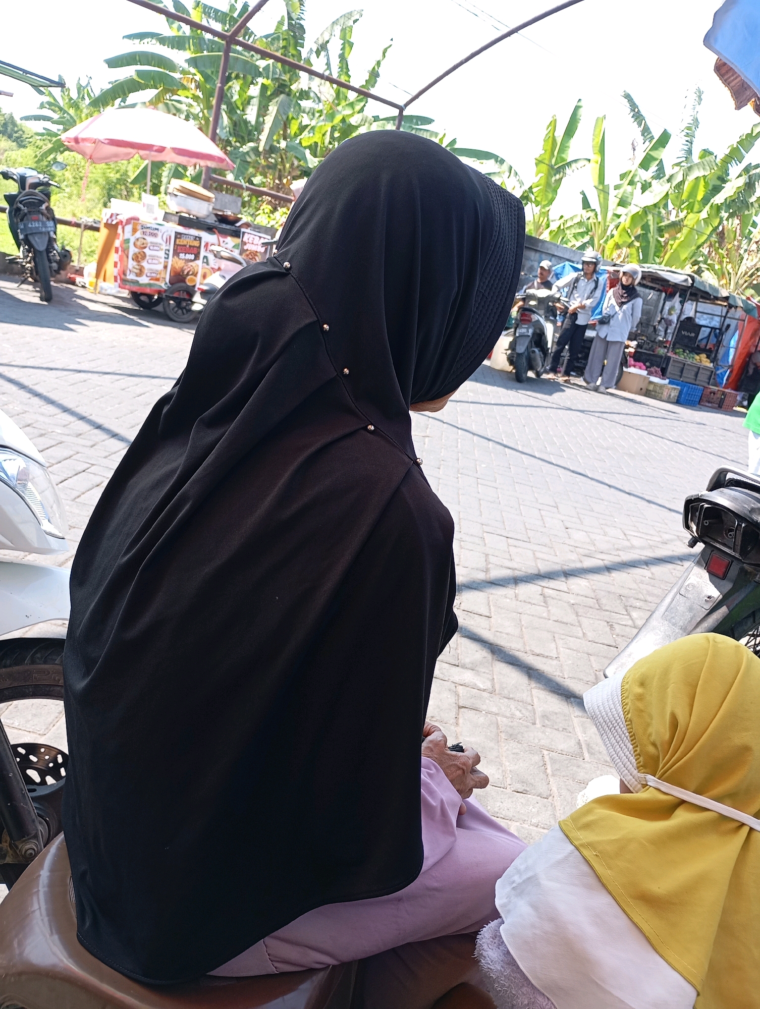 In the city of Kalisampurno, Indonesia, on a bright, sunny day, an outdoor scene unfolds with a focus on two individuals. The foreground is dominated by the back of a person wearing a black hijab and a light purple garment, seated on what appears to be a brown vehicle seat, possibly a motorcycle. Their hand is visible, holding a dark object. To the right, a second person is partially visible, wearing a yellow hijab with a white trim, also seated. Behind them, a paved walkway is visible, interspersed with shadows. In the mid-ground, a food cart is set up with a pink umbrella overhead. Various food items are displayed on the cart, with prices indicated in Indonesian Rupiah. Several people are gathered around the food cart and in the background. A motorcycle is parked to the left of the food cart. Further back, more people are seen, some wearing helmets, and a stall selling fruits is visible. The environment is characterized by lush green banana trees and other foliage, indicating a tropical setting. The overall atmosphere suggests a lively street market or a roadside food stall area.