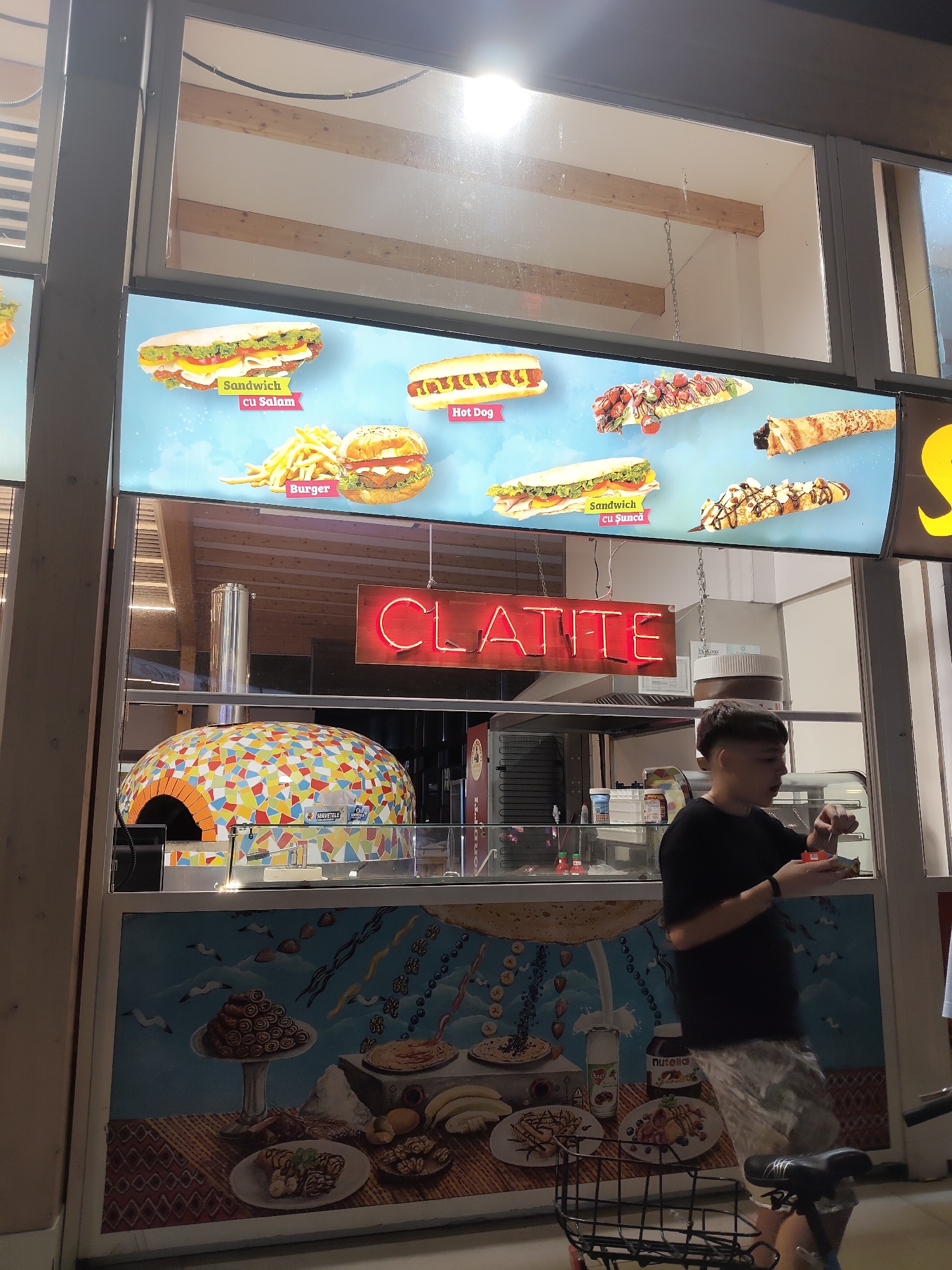 The image depicts an outdoor food stall, likely in Constanța, Romania, as indicated by the "CLATITE" sign, which translates to "pancakes" in Romanian. The stall features a colorful, mosaic-tiled pizza oven and a display window showcasing various food items like sandwiches, hot dogs, burgers, and crepes. A young person in a black t-shirt and patterned shorts is seen interacting with food items behind the counter. The stall has a menu board above the display window with images and text in Romanian. The overall setting appears to be a street or promenade, with a bicycle parked in the foreground. The lighting suggests it is evening or nighttime.