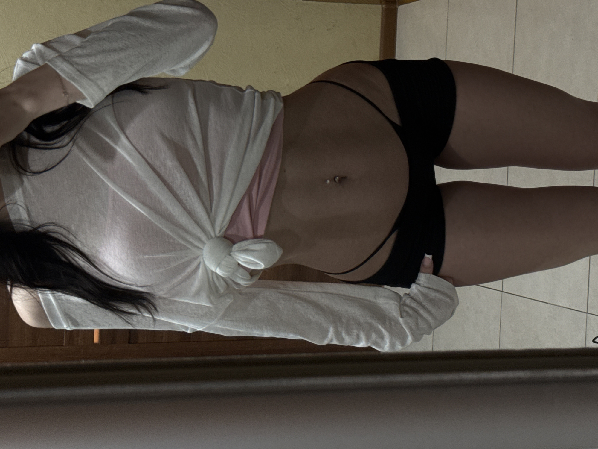 The image shows a person's midsection in a room. The person is wearing a white long-sleeved shirt tied at the waist, a pink camisole underneath, and black shorts. They have a belly button piercing. The background consists of a tiled floor and a wall. The overall scene suggests an indoor setting, possibly a bedroom or dressing area. There is no visible indication of the city of Khimki, Russia.
