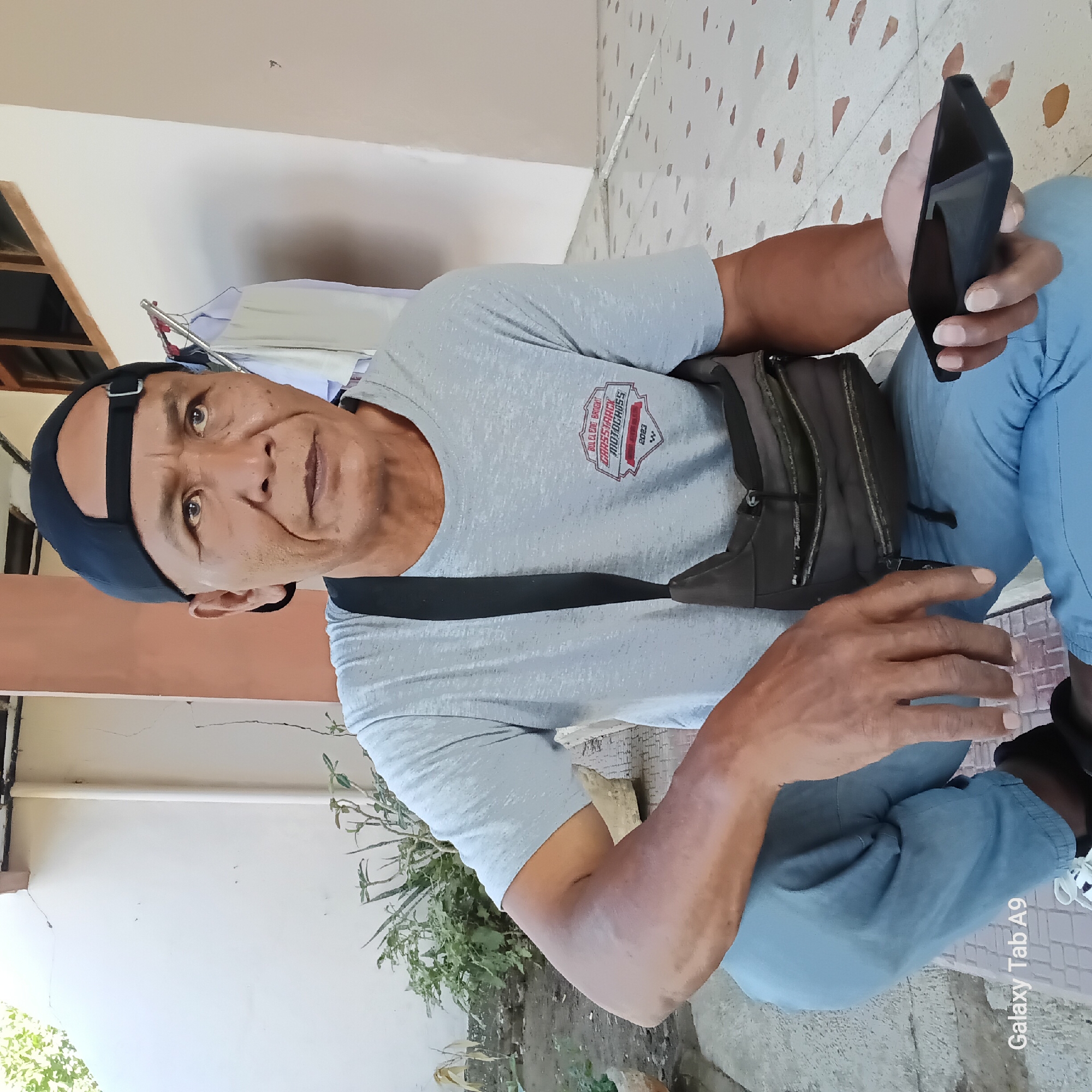 The image shows a middle-aged man squatting on a tiled patio in front of a building, possibly in Sambirejo, Indonesia, given the text overlay "Galaxy Tab A9" which is a common device in the region. The man is wearing a grey t-shirt with a logo, a black baseball cap, and light blue pants. He has a dark-colored sling bag across his chest. In his right hand, he is holding a dark-colored smartphone. The background consists of a light-colored wall, some potted plants, and laundry hanging on a rack. The overall scene appears to be an outdoor residential or communal area.