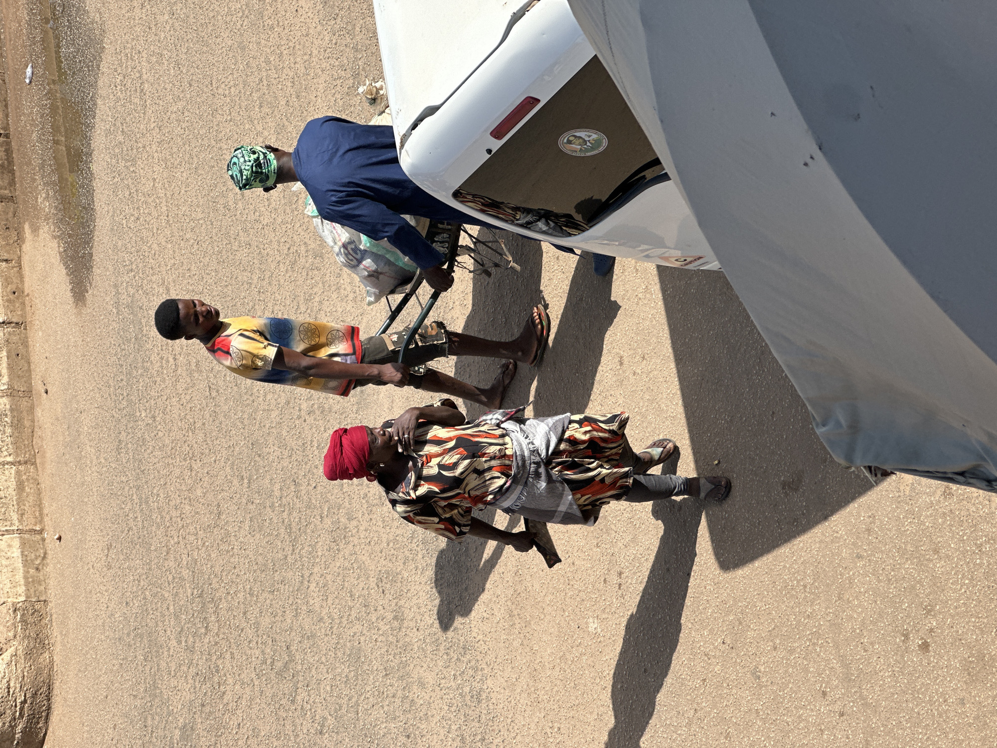 In Bukuru, Nigeria, a street scene unfolds with three individuals. In the foreground, a person wearing a vibrant red headscarf and a patterned dress walks towards the left, holding a dark object in their right hand. To their right, a man in a colorful t-shirt and shorts looks towards the man pushing a cart. The man pushing the cart, clad in a dark blue robe and a patterned green cap, is engaged in moving a load of bagged goods. The ground is paved with asphalt, showing signs of wear. The environment is illuminated by natural sunlight, casting distinct shadows. A white vehicle, partially visible on the right, dominates the lower right quadrant of the frame. A faint shadow is cast on the ground by the central figures.