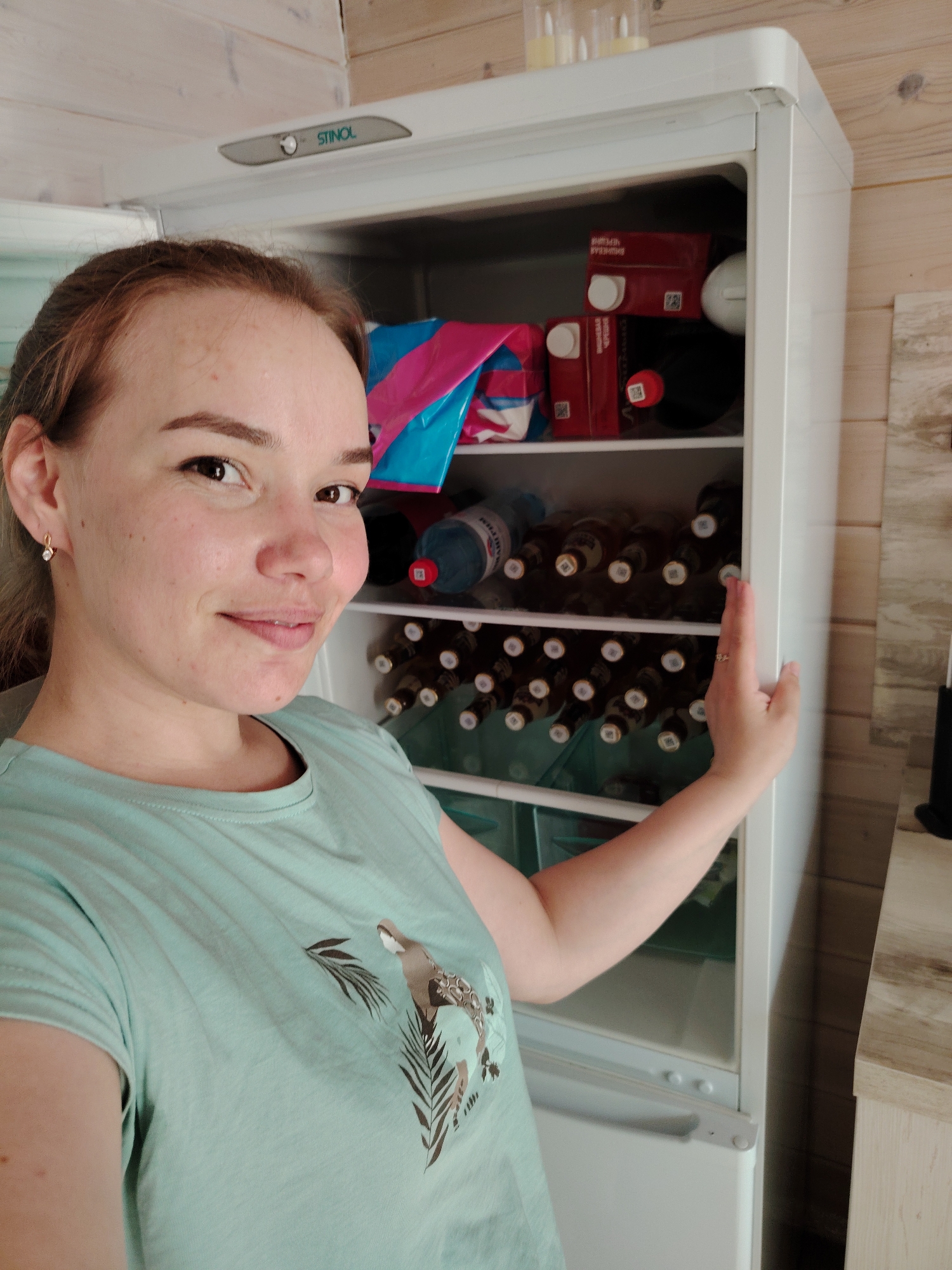 A woman is holding open the door of a white Stinol refrigerator in a room with light-colored wooden paneling. The shelves of the refrigerator are filled with bottles, possibly beer or wine, along with a couple of juice boxes and a colorful bag. The woman, who appears to be in her 20s or 30s, is wearing a light blue-green t-shirt with a nature-themed graphic and is looking directly at the camera with a slight smile. This image was taken in Malyi Bukor, Russia.