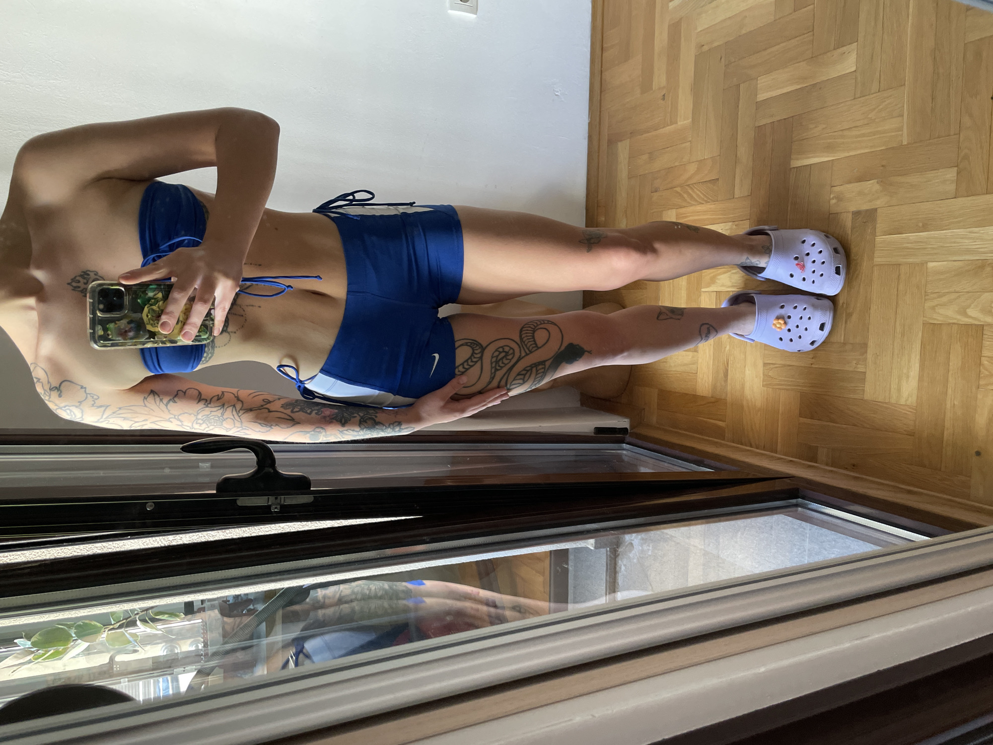 The image captures a full-body mirror selfie of a young adult female standing indoors, located in Boreti, Montenegro. She exhibits a slender, athletic build and is dressed in a royal blue two-piece outfit, consisting of a strapless bandeau top with central drawstrings and high-cut shorts featuring white vertical stripes on the sides and side drawstrings. A white Nike swoosh logo is visible on the left thigh of the shorts.
The individual displays extensive body art, including floral or foliage tattoos covering both her right and left arms and shoulders. Intricate patterns are tattooed on her sternum and upper abdomen, partially obscured by the smartphone she holds in her right hand. A large, coiled snake tattoo is prominently featured on her right thigh, and smaller, scattered tattoos are visible on her lower legs, including one resembling a butterfly. She is wearing light purple Crocs clogs; the left clog is accessorized with a small yellow-orange flower charm, and the right clog with a pink charm.
The background to the right features a plain white wall with a visible white electrical outlet. To the left, a large window or glass door with dark brown frames reflects the subject and the interior. Through the window, an outdoor scene is partially visible, including a green potted plant with variegated leaves and an outdoor paved area. A black window handle is affixed to the frame. The floor is composed of light-colored wooden parquet flooring laid in a herringbone pattern. The scene is brightly illuminated by natural daylight.