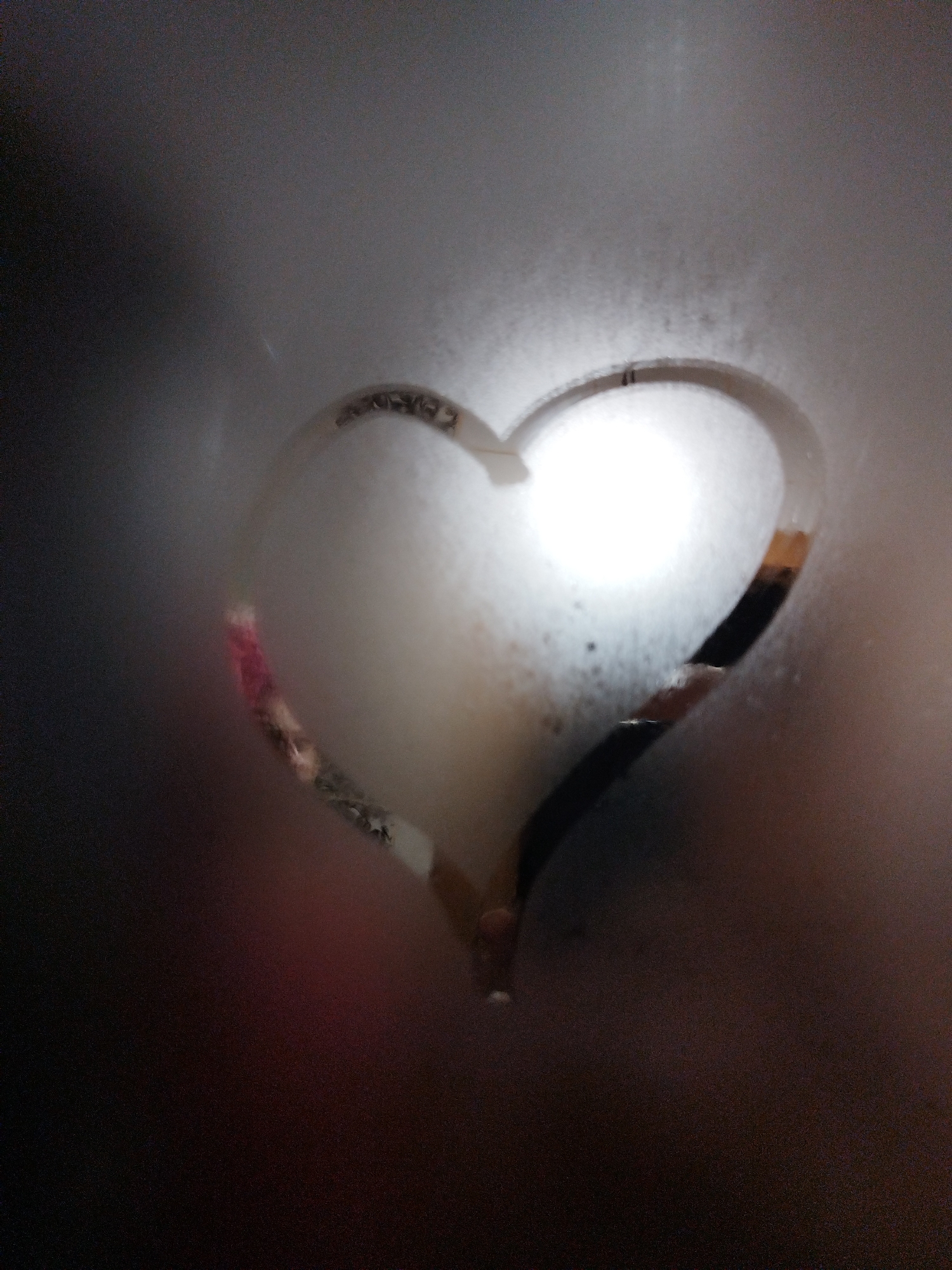A heart shape is drawn in condensation on a glass surface. The heart has a gradient of colors within its outline, ranging from pink to black. A bright light source illuminates the center of the heart, creating a halo effect. The background is dark and out of focus.