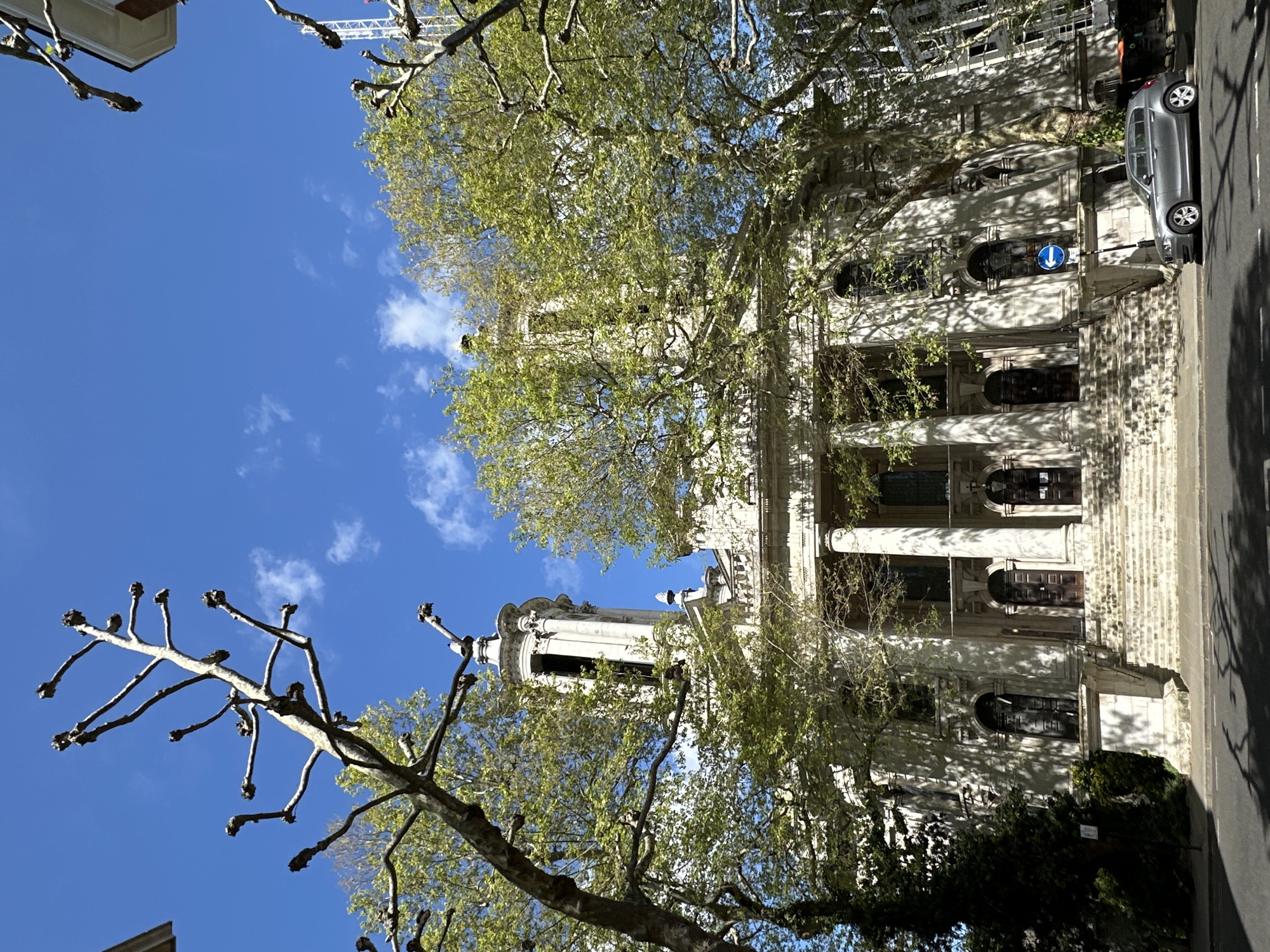 The image captures St. Marylebone Parish Church in London, United Kingdom. The architectural style is Neoclassical, with prominent Corinthian columns and a steeple. The church facade is made of light-colored stone. Mature trees with burgeoning leaves partially obscure the building. A street sign indicating traffic direction is visible adjacent to the church. A gray vehicle is parked along the roadside, and buildings line the street. The sky is blue with scattered clouds.