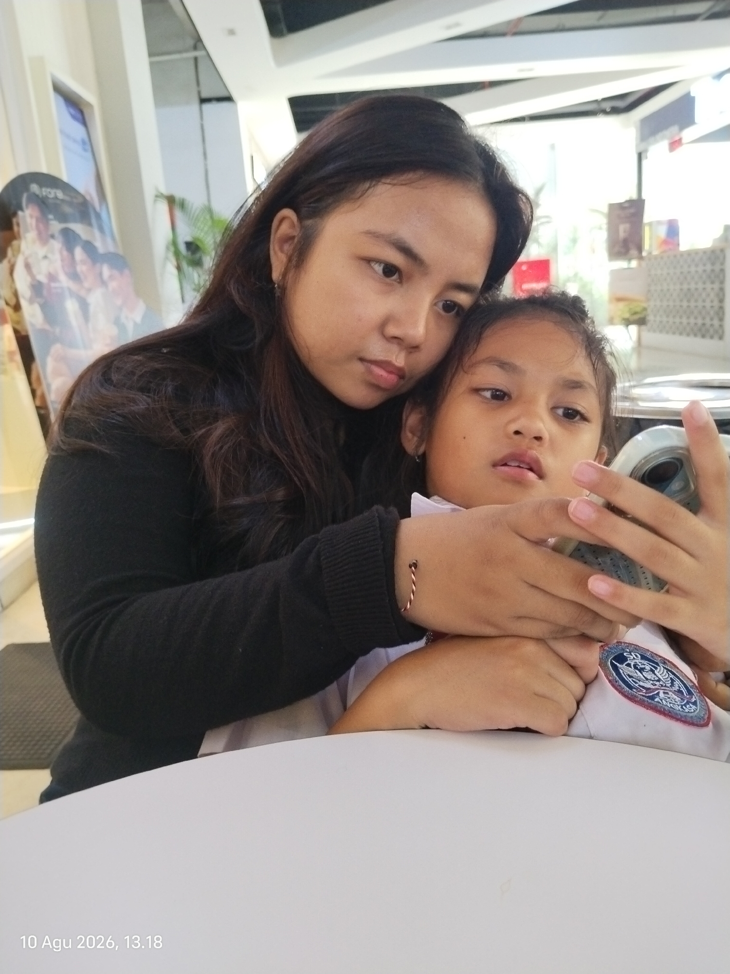 An overhead shot shows a mother and daughter sitting at a white table, likely in a restaurant or mall in East Jakarta, Indonesia, given the timestamp "10 Agu 2026, 13.18". The mother, with long dark hair and wearing a black sweater, is leaning in and holding a mobile phone, which she is showing to the daughter. The daughter, a young girl with dark hair and wearing a white collared shirt with a blue and red patch on the sleeve, is looking intently at the phone. Her hands are also holding the phone, suggesting she is actively engaged with what is on the screen. The background is blurred but shows a bright, modern interior with architectural elements and some signage, indicating a public space. The overall scene is intimate, focusing on the shared activity between mother and child.