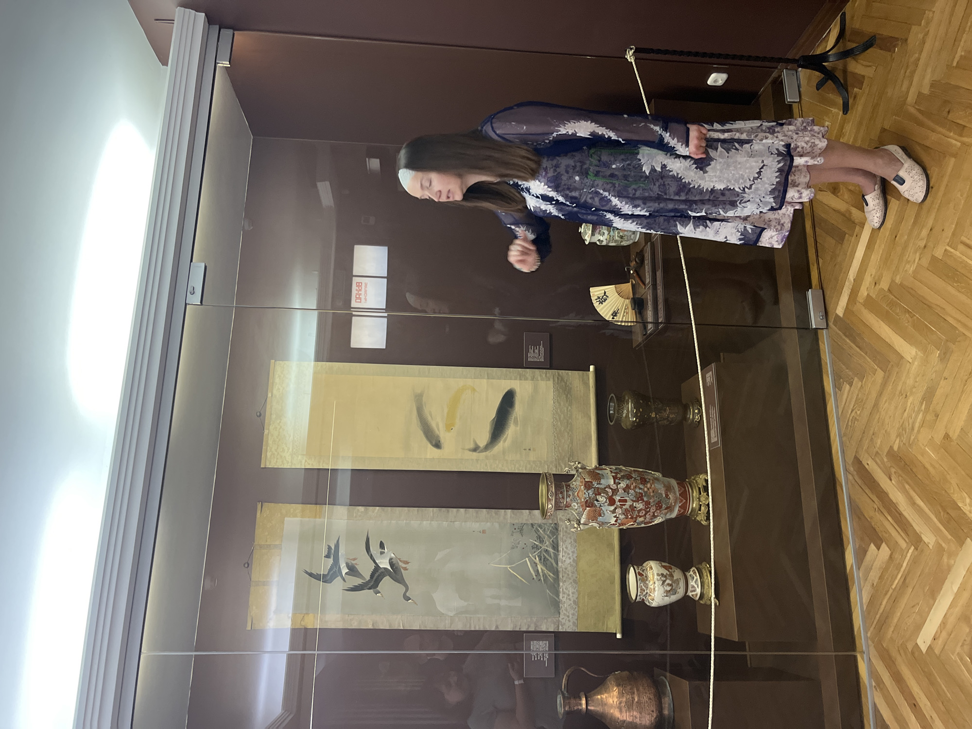 A woman, wearing a blue and white patterned dress and light-colored shoes, stands in front of a glass display case in a museum. She is gesturing with her right hand. Inside the display case are several items, including two scrolls with paintings of birds and fish, a large ornate vase, two smaller vases, and a fan. To the left of the display case, a copper pitcher is visible. The floor is a herringbone pattern of light-colored wood. The museum appears to be the Brest City Museum of Local Lore in Brest, Belarus, based on the presence of historical artifacts typical of such institutions.