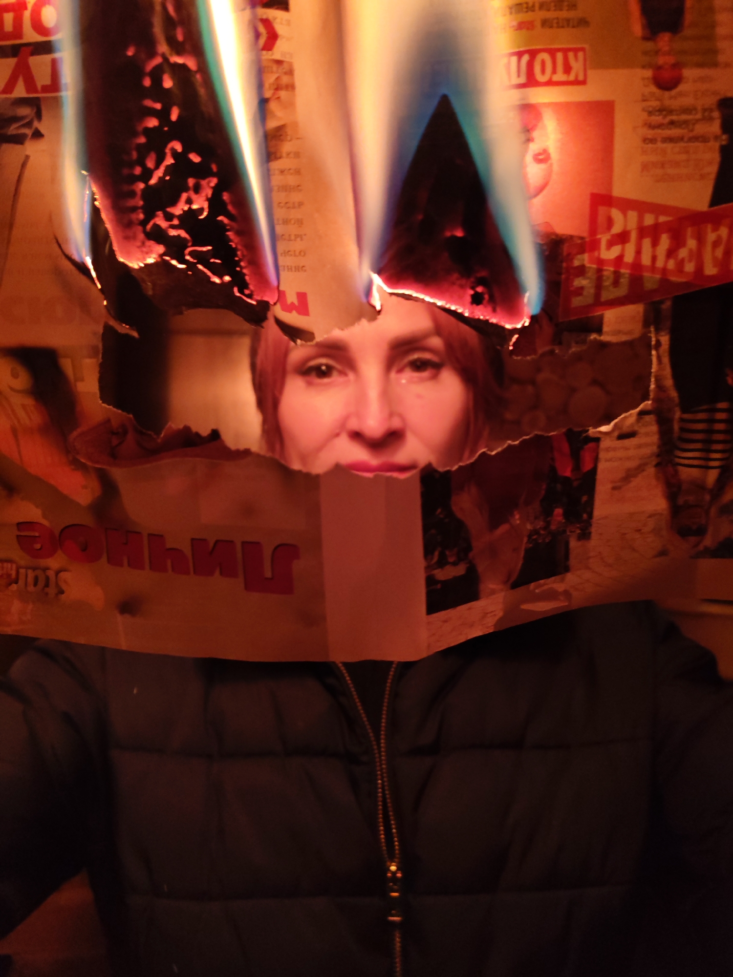 A woman stands behind a burning, torn newspaper. The flames are visible above her head, with the charred edges of the paper surrounding her face. The newspaper's text includes Russian words like "Личное". The background behind the newspaper consists of various posters and images. The woman is wearing a dark puffer jacket with a gold zipper. The setting appears to be indoors.