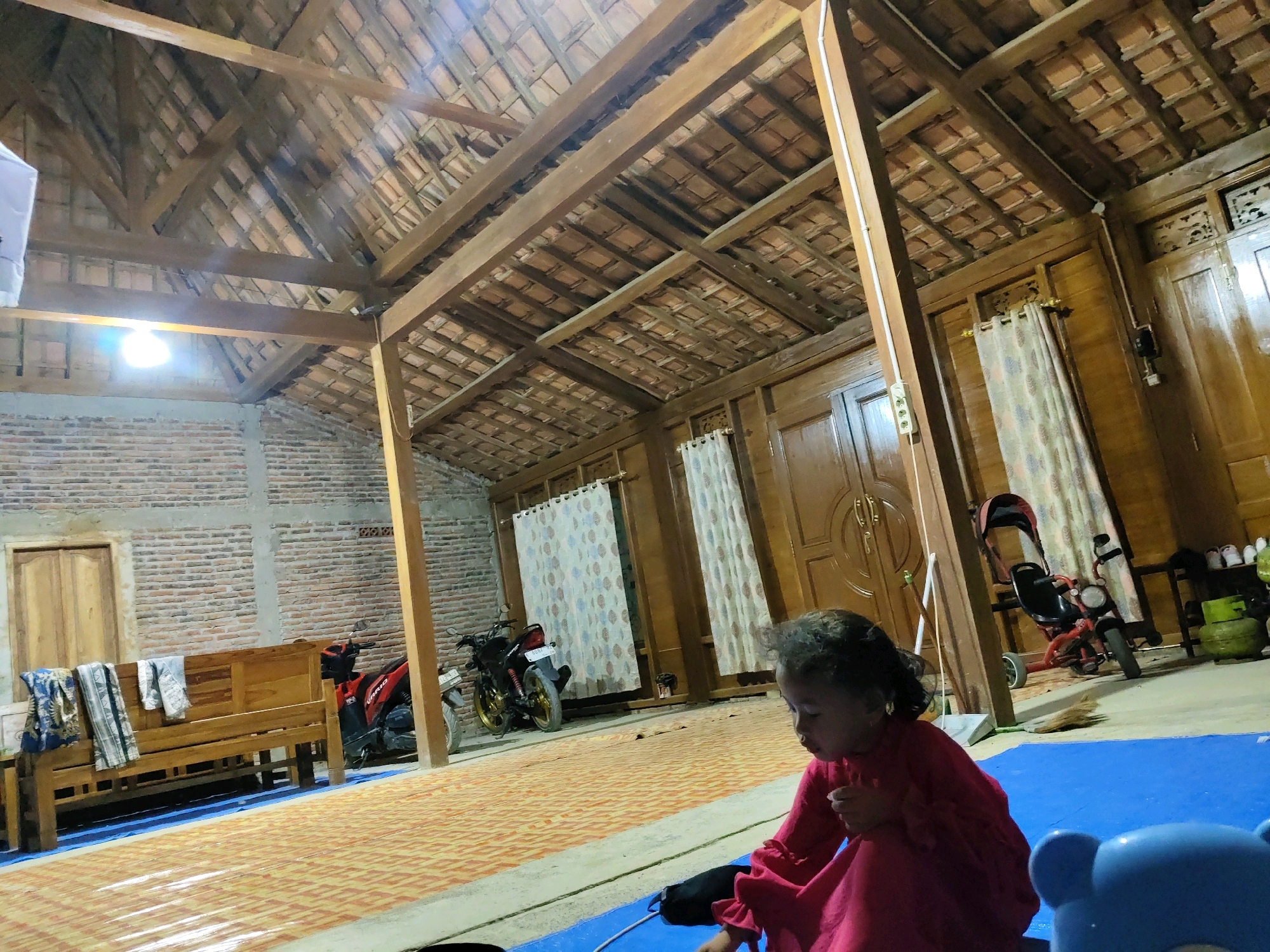 A young girl in a pink dress is seated on a blue mat inside a wooden structure in Wirosari, Indonesia. The structure appears to be an open-air dwelling or workshop with a tiled roof supported by wooden beams. To the left, a wooden bench is placed against a brick wall, with folded textiles resting on it. Two motorcycles are parked nearby. The girl is looking down at something in her hands. In the background, there are wooden doors, curtains, and more wooden structures. The scene is illuminated by artificial light.