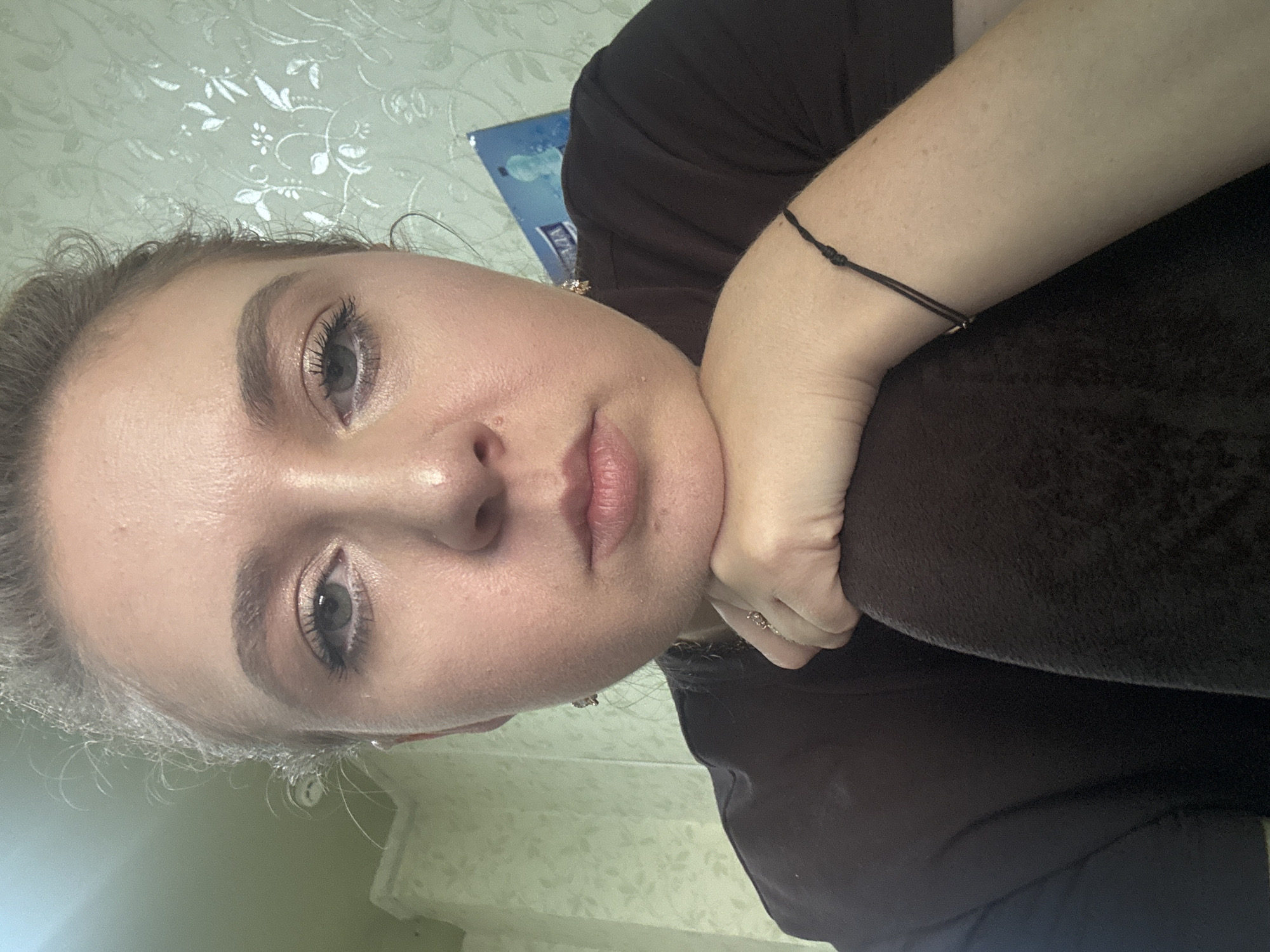 A close-up, selfie-style, medium shot depicts a young woman with light skin and light brown hair pulled back into a bun. She is wearing dark brown clothing and a black string bracelet on her left wrist. Her chin rests on her left hand, which is positioned on her right arm. Her eyes are open and looking at the camera. The background consists of a light green, textured wallpaper with a floral pattern and a blue and white poster. The lighting is soft and diffused, suggesting an indoor environment. There is no indication of the city or specific location within the image.