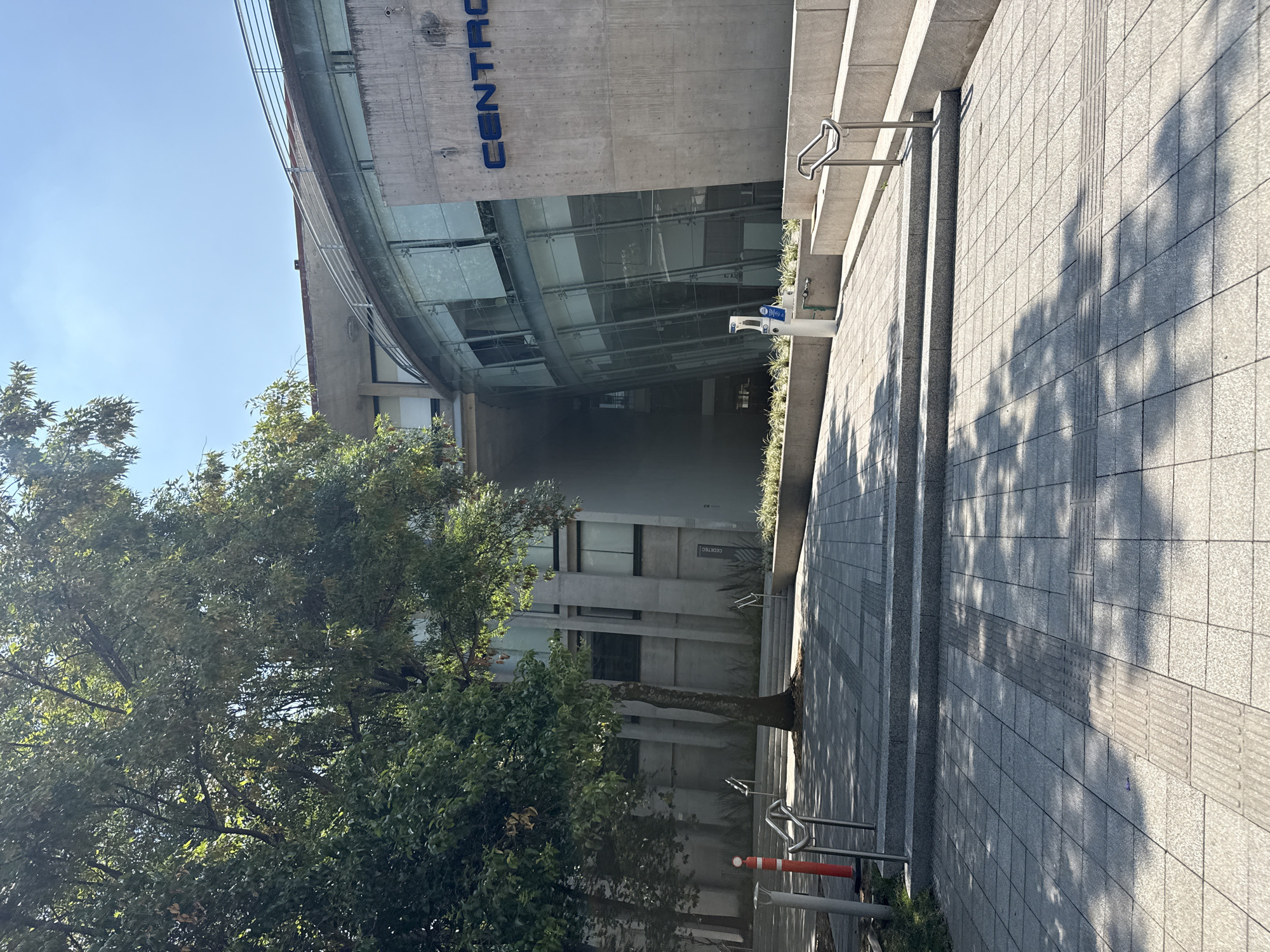 The image shows an exterior view of a building labeled "CENTRO" in Mexico City, Mexico. The building features a concrete facade with a curved glass window section. In front of the building is a tree with dense foliage, casting shadows on the paved walkway leading to the building's entrance. A set of stairs with a handrail is visible along the right side of the image. There are bike racks near the base of the stairs, with a few orange and white elements. The sky is clear and blue. The name of the building, "CEDETEC," can also be observed, although it is partially obscured.